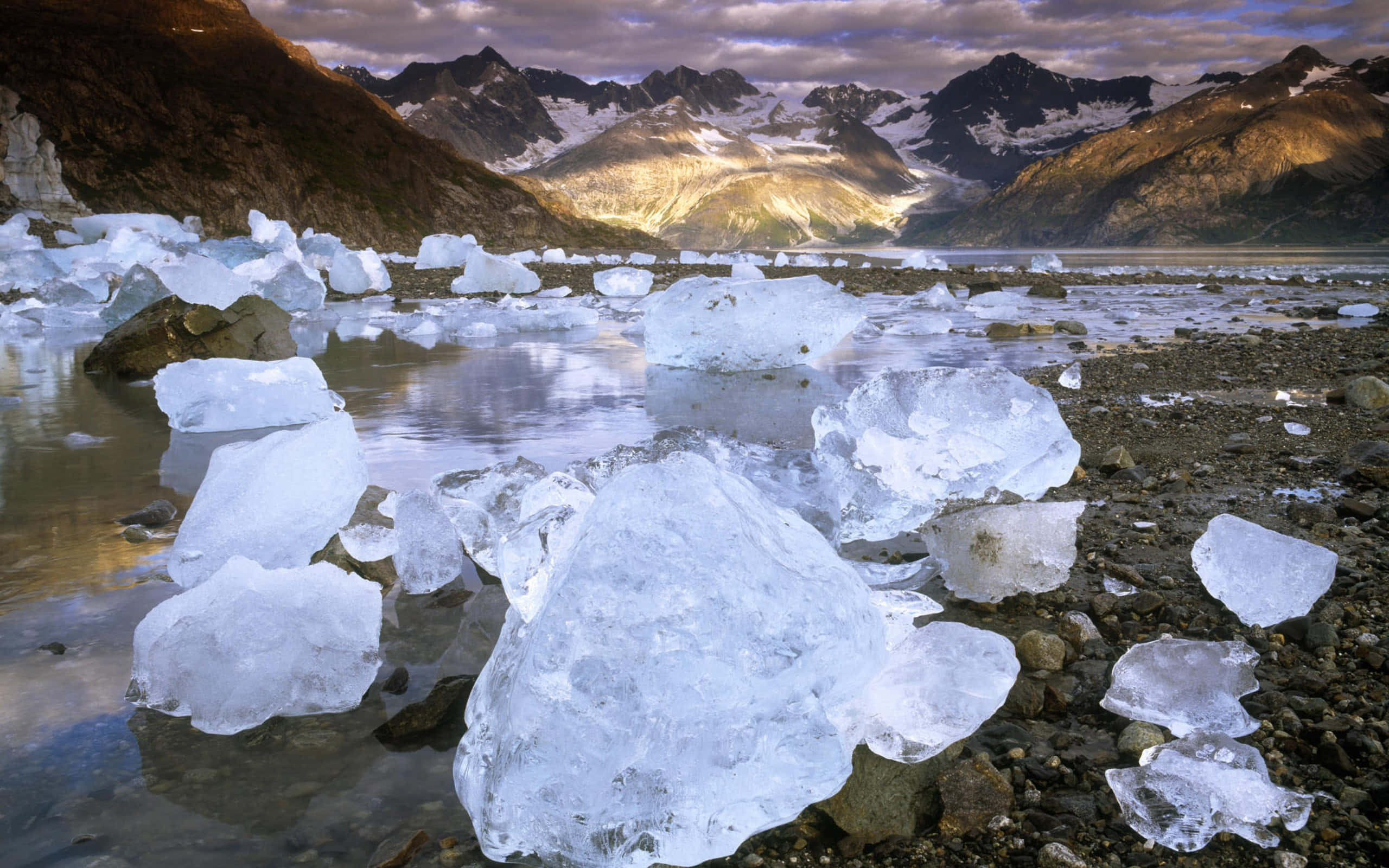 冰 桌布 壁紙 — 閃閃發光的冰山漂浮在冰川湖中，周圍是白雪覆蓋的山脈，天空呈現戲劇性的暮色。