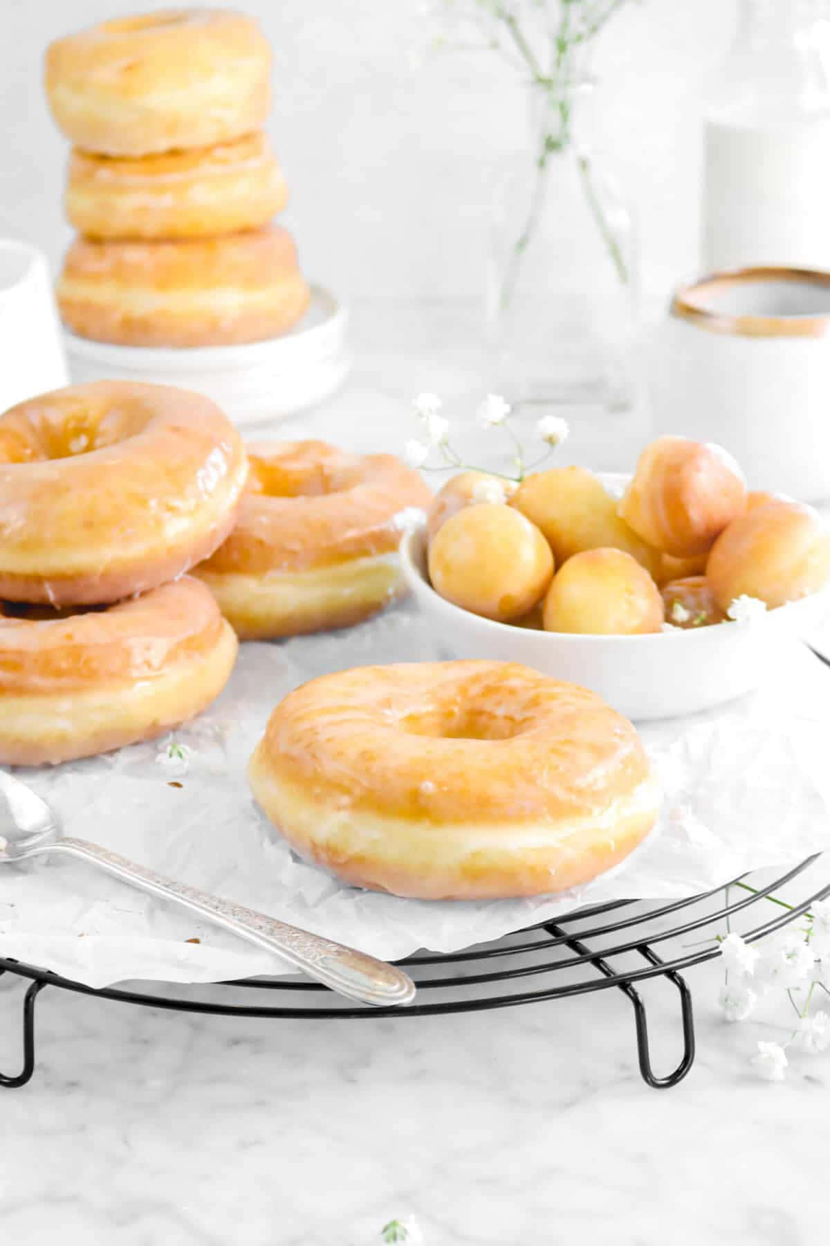 Glazed Donut 桌布 壁紙 — 閃閃發光的釉面甜甜圈和金色迷你甜甜圈放在金屬架上，旁邊有一束滿天星花瓶和一個銀勺，置於大理石檯面上。