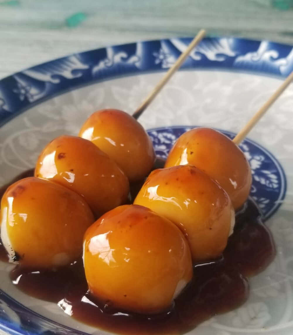 Japanese Sweets 桌布 壁紙 — 閃閃發光的金黃色餃子串放在深色醬汁中，置於藍白圖案盤上。
