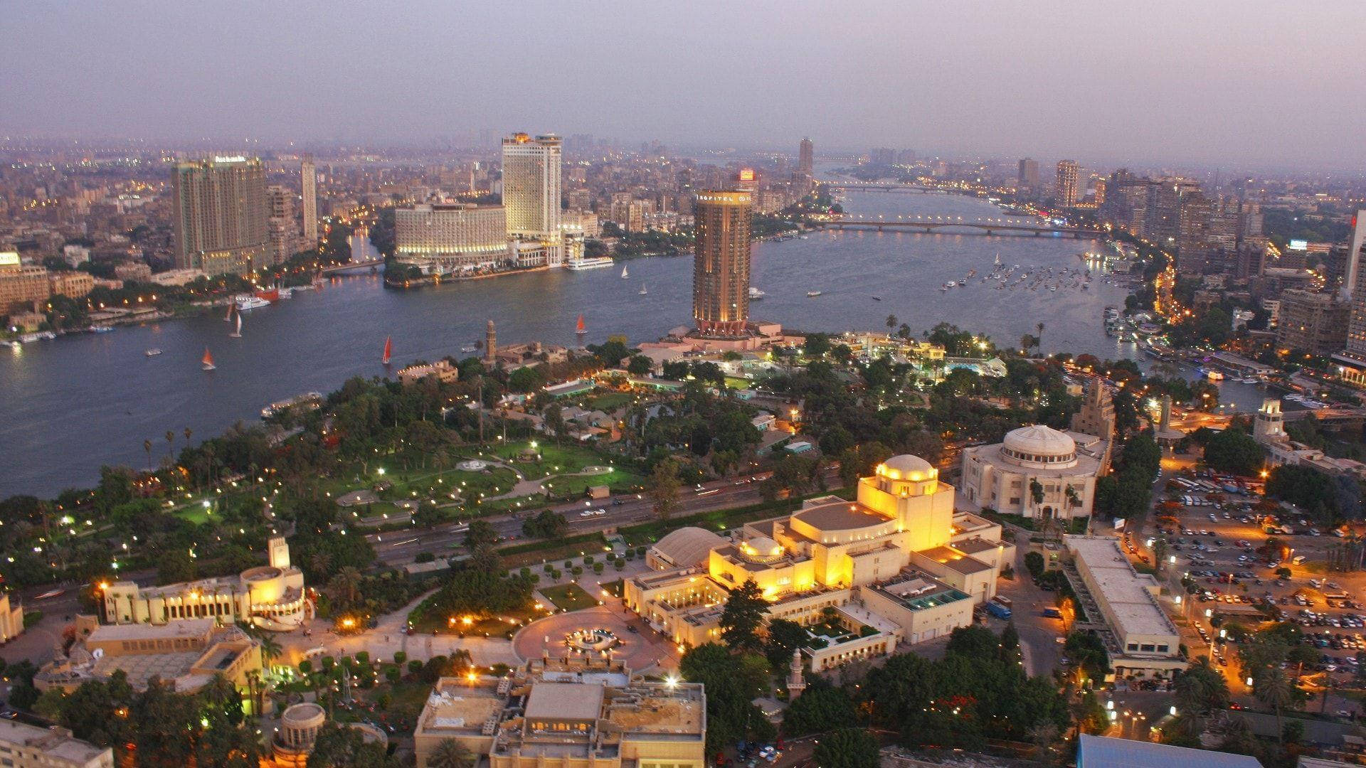 Cairo 桌布 壁紙 — 開羅黃昏時的鳥瞰圖，尼羅河在城市燈光下閃耀，歷史建築在柔和的紫色天空映襯下亮起。