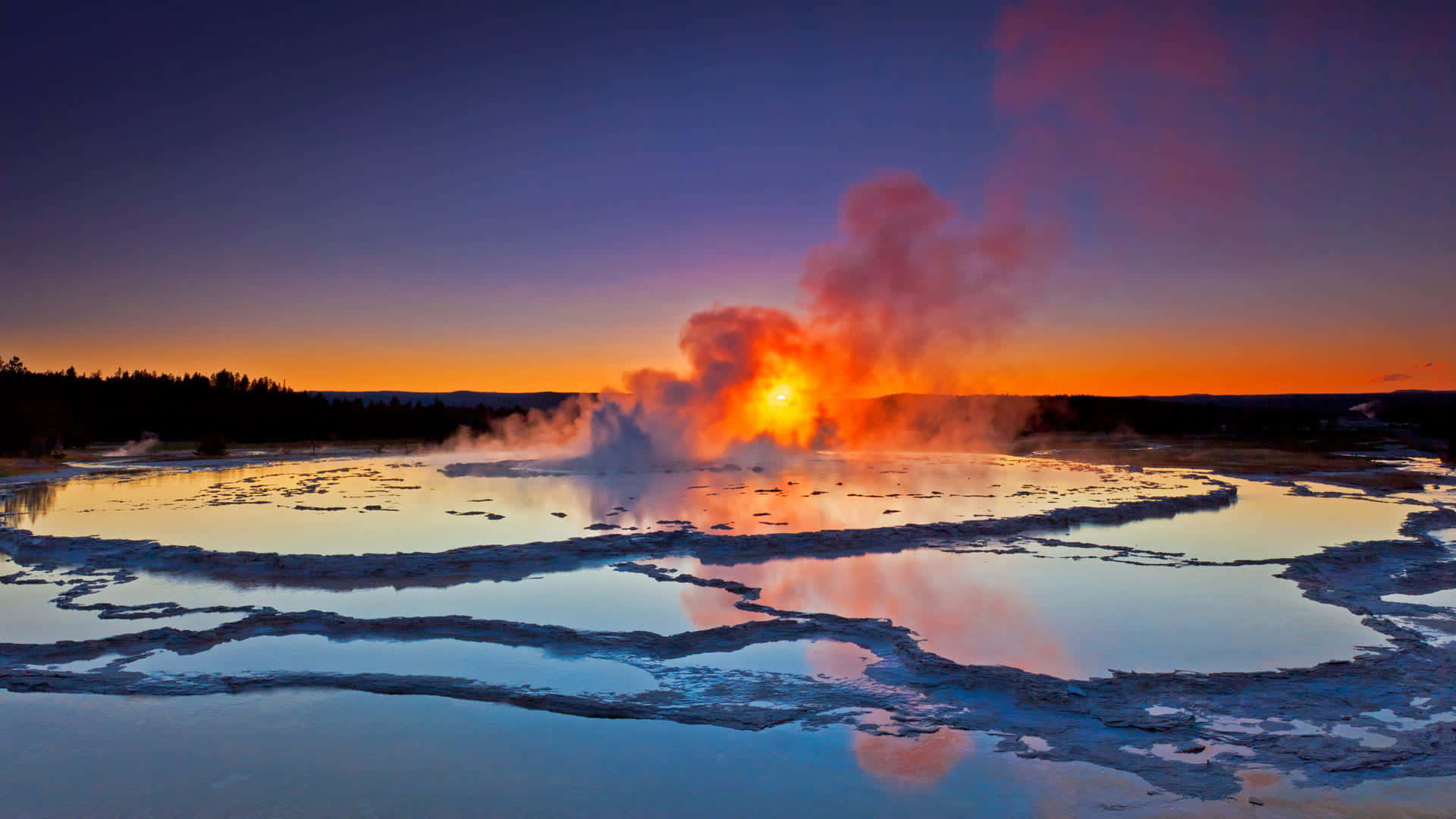Yellowstone Geysers 桌布 壁紙 — 間歇泉在暮色天空下以火熱橙色噴發，倒影映在冰冷的溫泉池中。