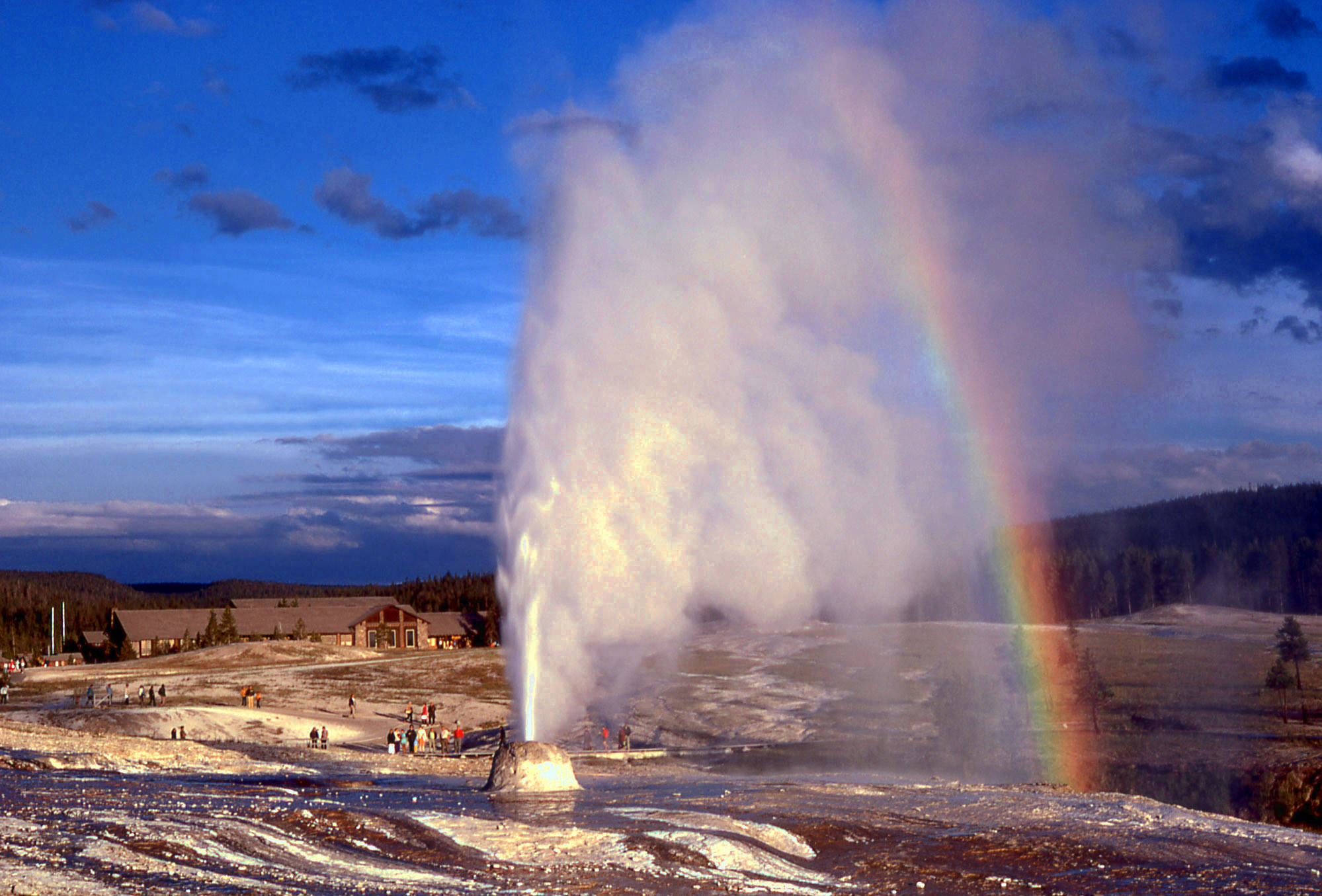 Yellowstone 桌布 壁紙 — 間歇泉在黃昏時戲劇性地噴發，在深藍色天空的映襯下，透過蒸汽形成一道鮮豔的彩虹。