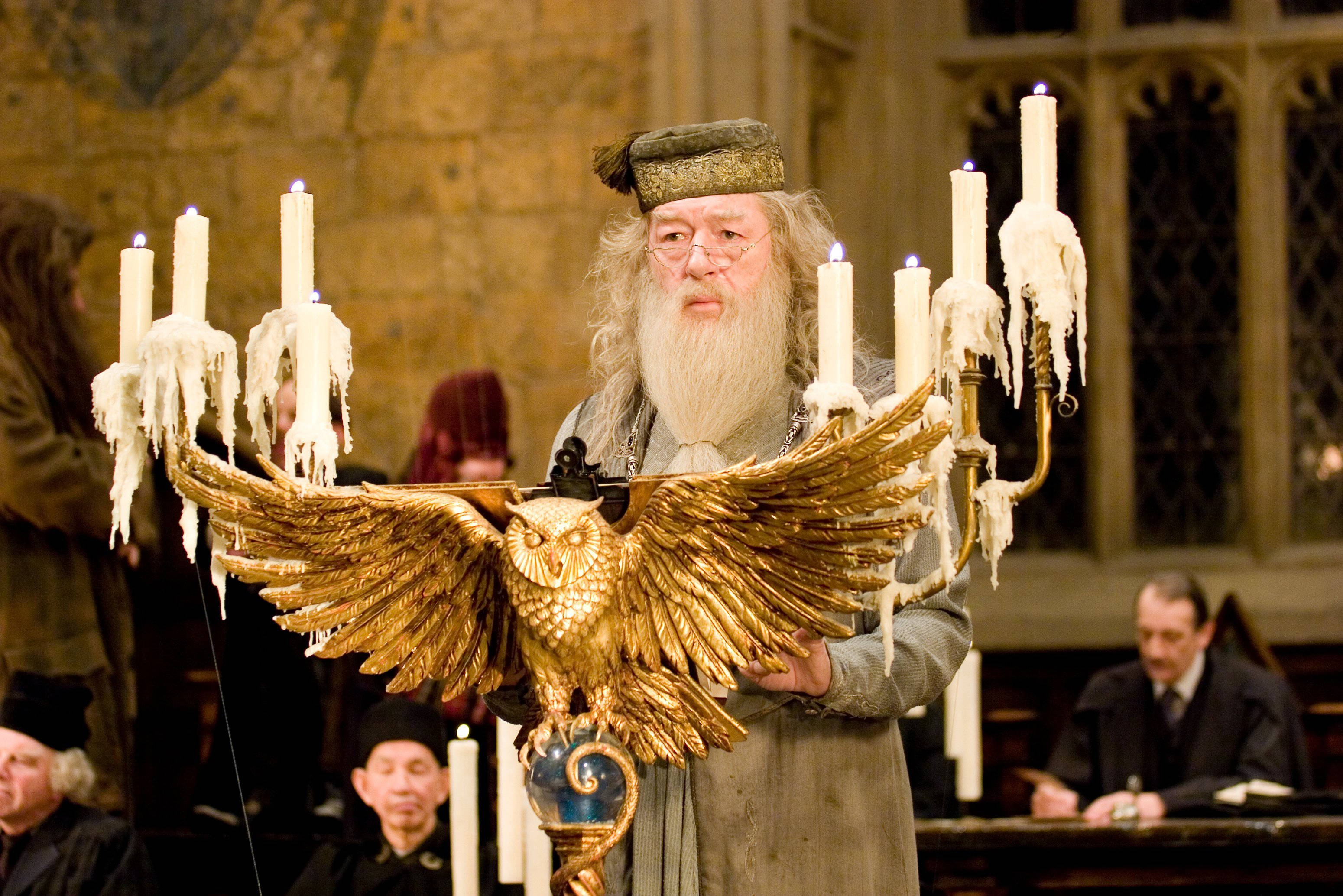 dumbledore 桌布 壁紙 — 阿不思·鄧不利多在一個宏偉的石廳中手持一個金色的貓頭鷹形燭台，燭台上插著正在融化的蠟燭。