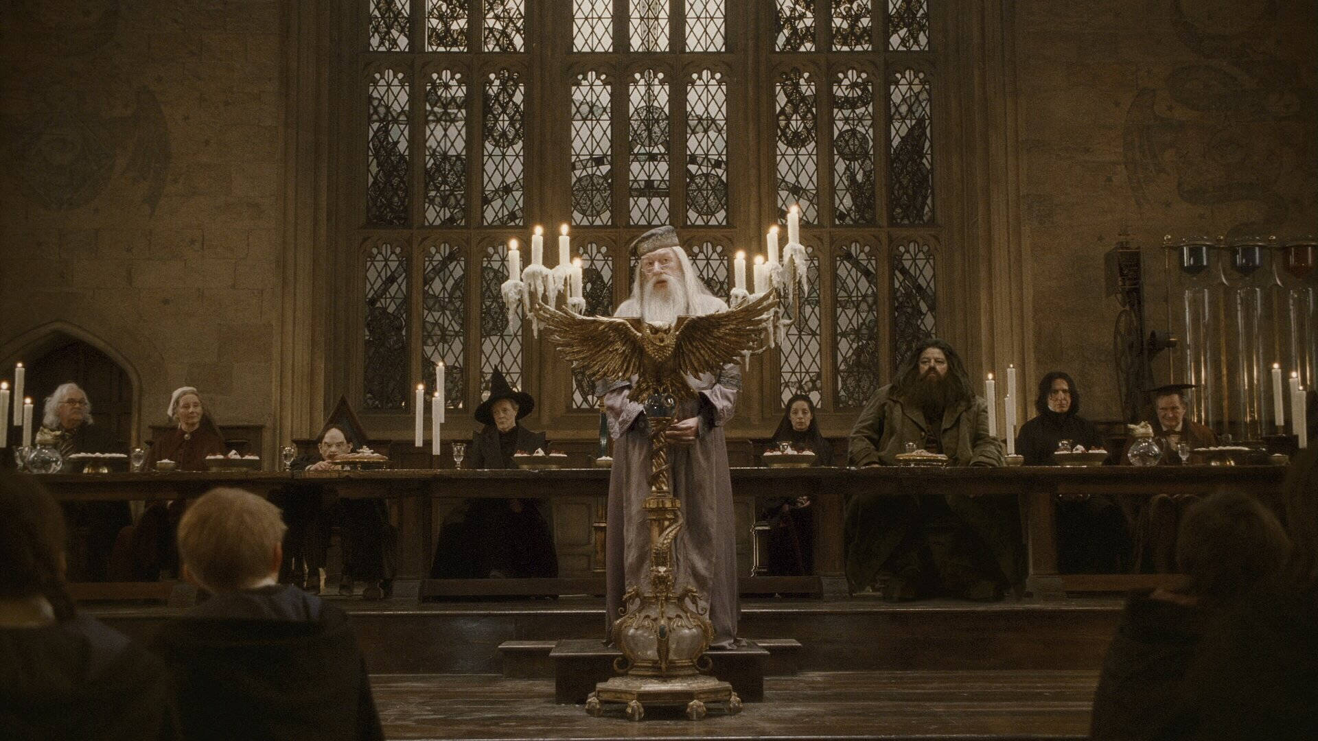 rubeus hagrid 桌布 壁紙 — 阿不思·鄧不利多莊嚴地站在一個宏偉的石廳中的金色講台上，廳內有燭光和彩繪玻璃窗照明。