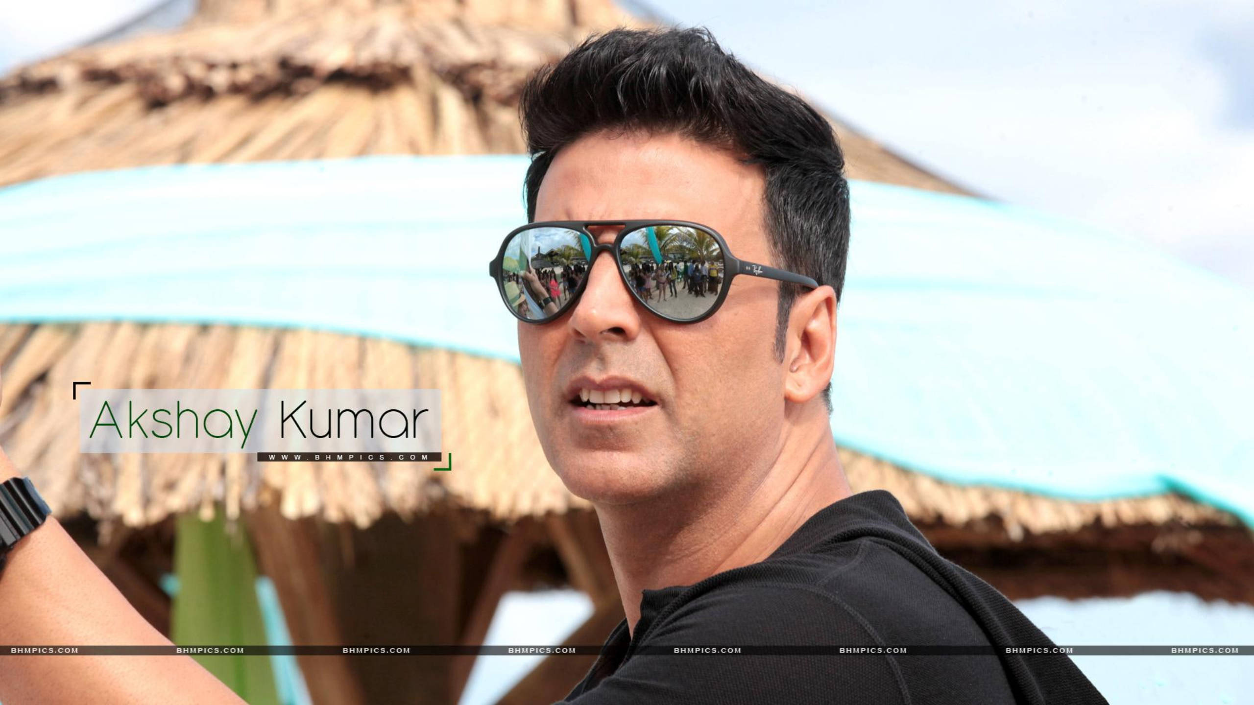 Akshay Kumar 桌布 壁紙 — 阿克謝·庫瑪爾戴著太陽眼鏡，在陽光明媚的海灘度假村茅草傘下自信微笑。