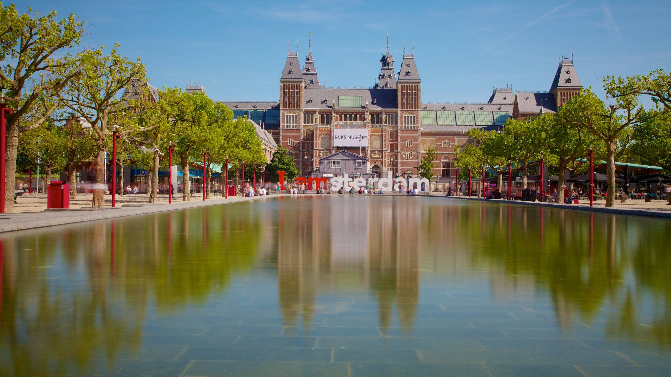 Netherlands 桌布 壁紙 — 阿姆斯特丹宏偉的國立博物館倒映在平靜的水景中，上方是明亮的藍天和茂密的綠樹。