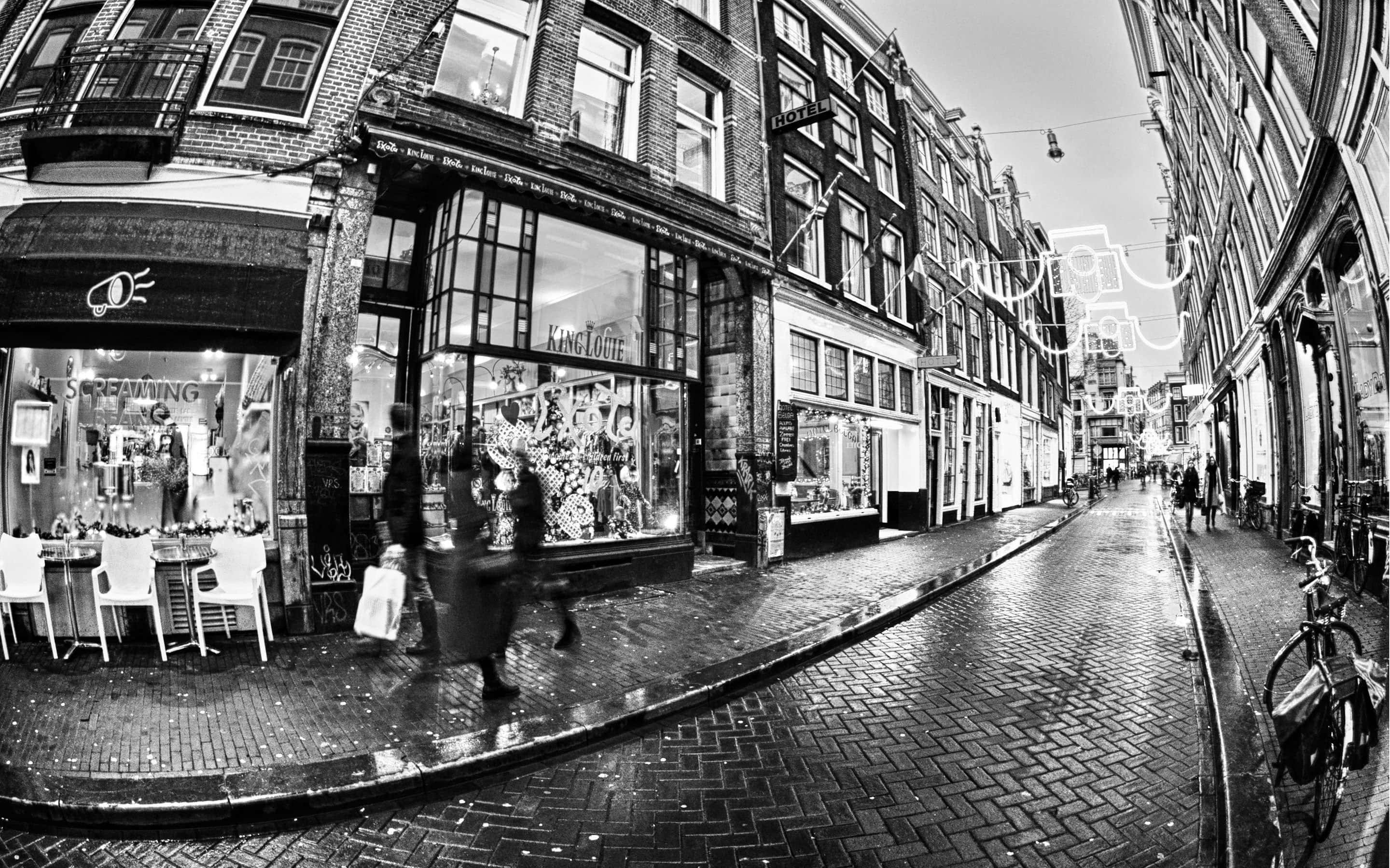 Black And White Architecture 桌布 壁紙 — 阿姆斯特丹濕滑鵝卵石街道的黑白魚眼鏡頭景觀，帶有模糊的行人和商店門面。