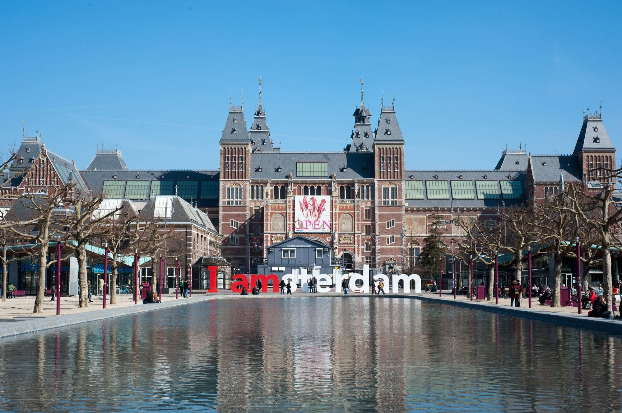 Netherlands 桌布 壁紙 — 阿姆斯特丹的宏偉國立博物館在清澈藍天下倒映在平靜的水景中，廣場兩旁排列著光禿的樹木。