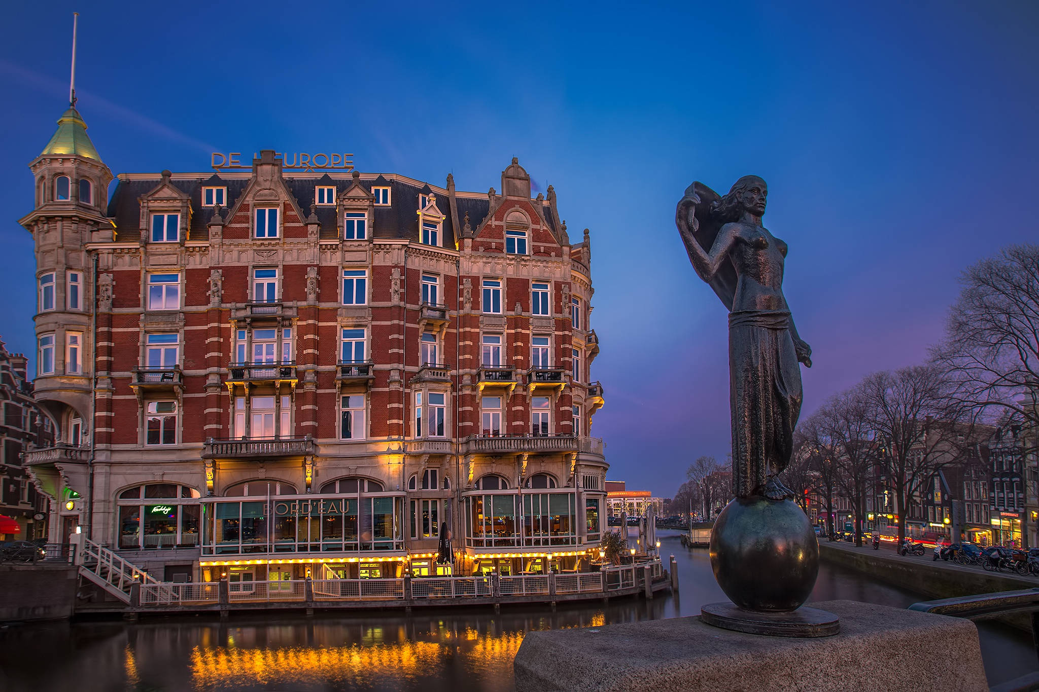 Netherlands 桌布 壁紙 — 阿姆斯特丹的暮色運河場景，一個球體上的銅像位於歷史悠久的紅磚建築旁，溫暖的燈光倒映在水中。