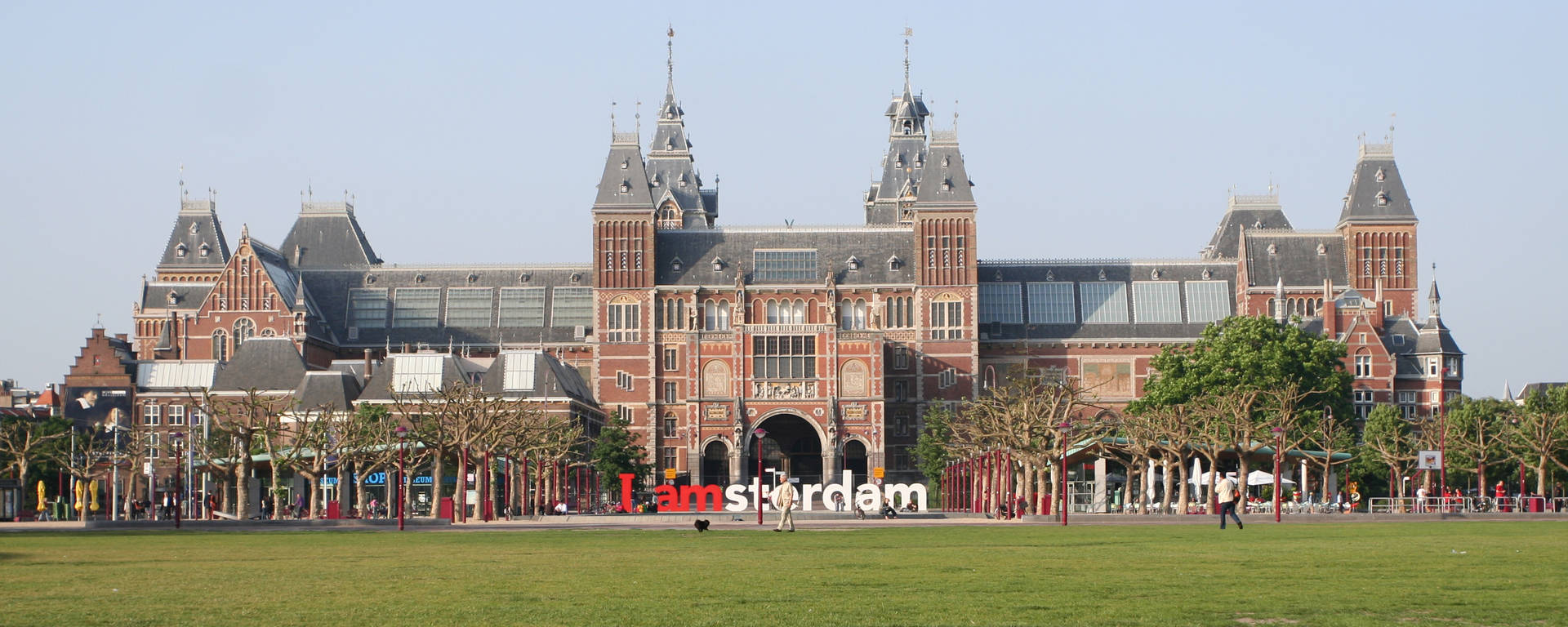 Museum 桌布 壁紙 — 阿姆斯特丹的紅色磚造國家博物館雄偉地矗立在充滿活力的綠色草坪後方，草坪上有「I amsterdam」標誌，人們在晴朗的藍天下漫步。