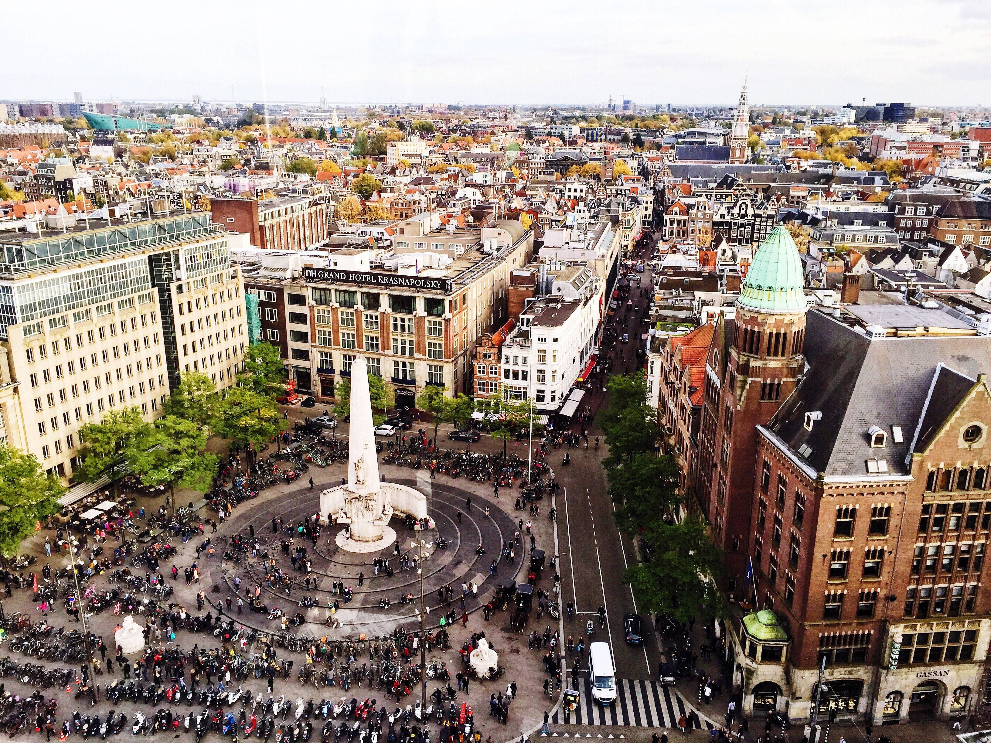 Amsterdam 桌布 壁紙 — 阿姆斯特丹繁忙廣場的鳥瞰圖，廣場上擠滿了人，聚集在中央紀念碑周圍，周圍環繞著歷史建築和秋樹。