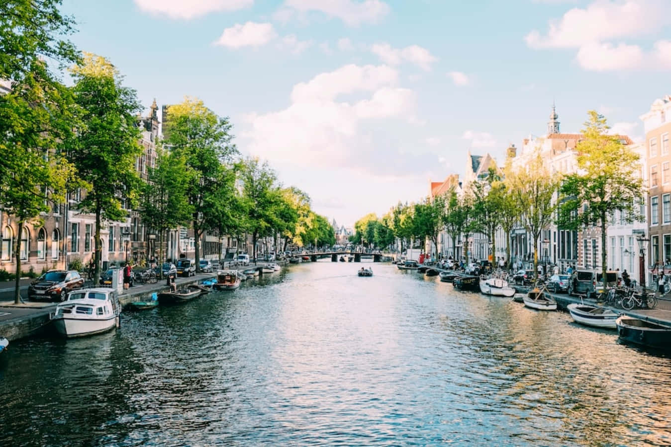 荷蘭 桌布 壁紙 — 阿姆斯特丹運河上晴朗的一天，歷史建築、樹木和船隻在部分多雲的天空下倒映在平靜的水面上。
