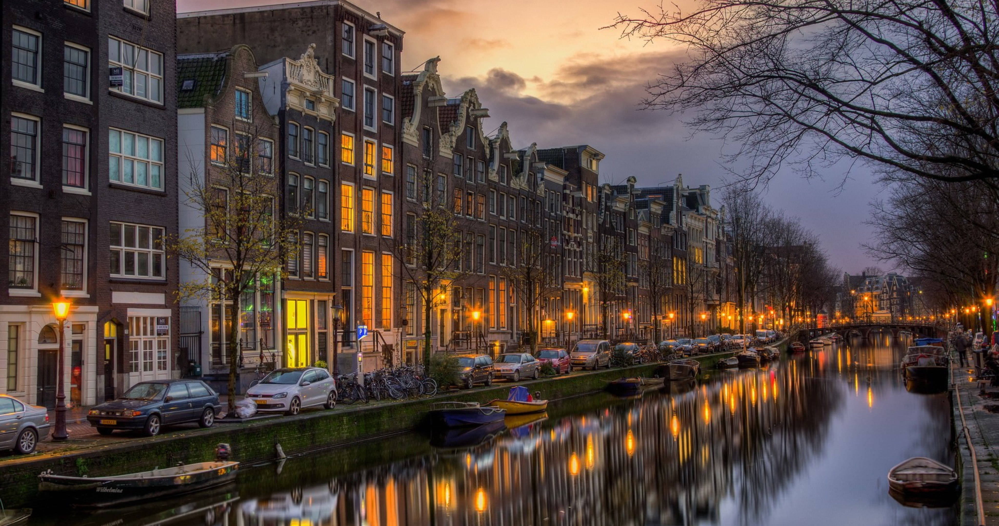 Amsterdam 桌布 壁紙 — 阿姆斯特丹運河在黃昏時分，溫暖的金色燈光倒映在平靜的水面上，旁邊是歷史悠久的山牆建築。