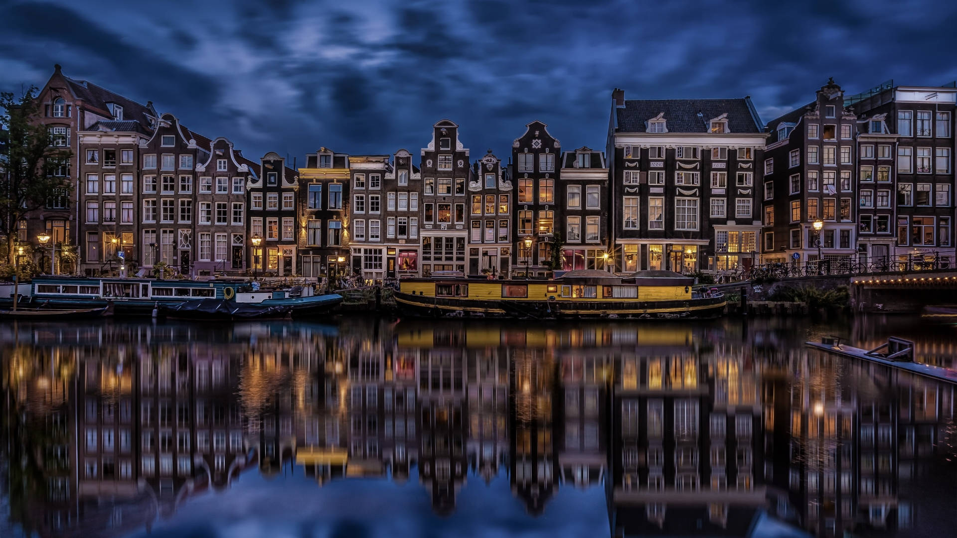 Netherlands 桌布 壁紙 — 阿姆斯特丹運河的寧靜黃昏景色，歷史悠久的山形建築和船隻倒映在平靜的水面上，天空憂鬱的藍色。
