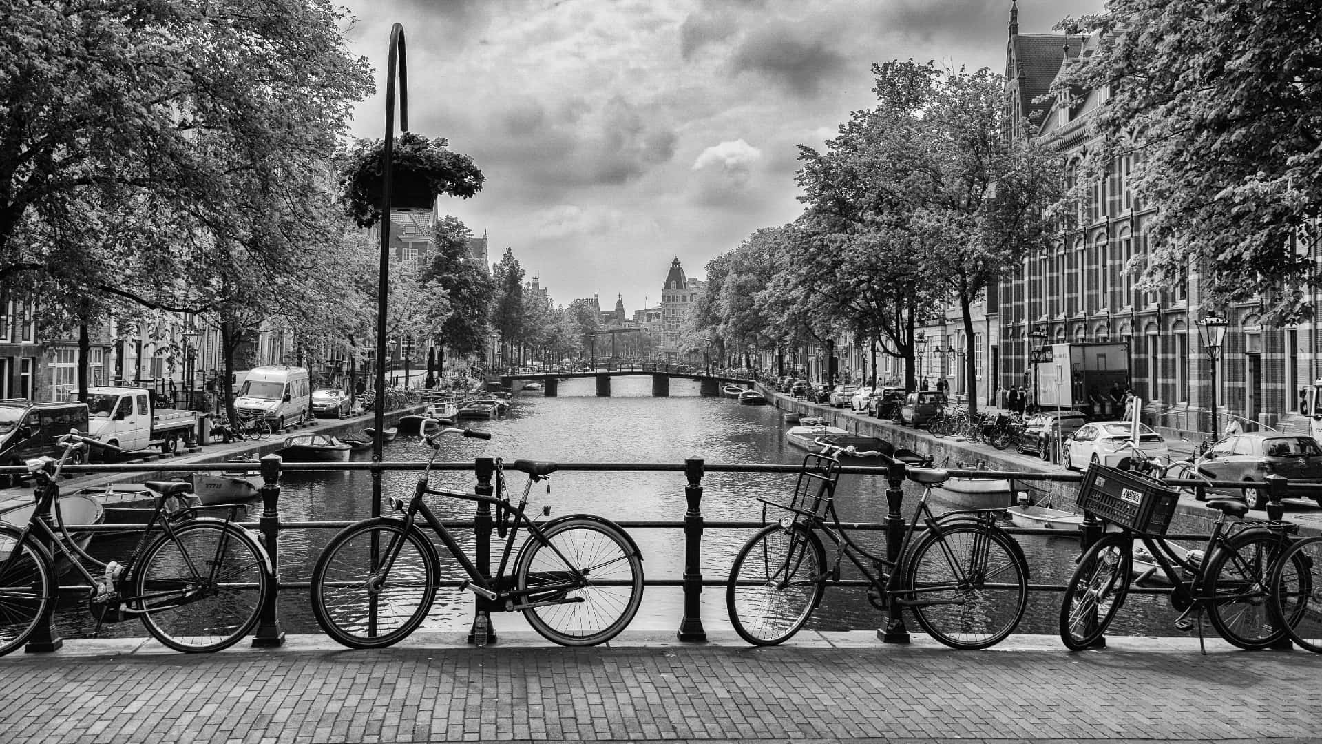 Black And White Cityscape 桌布 壁紙 — 阿姆斯特丹運河的黑白景觀，自行車沿著磚砌小路排列，天空多雲。