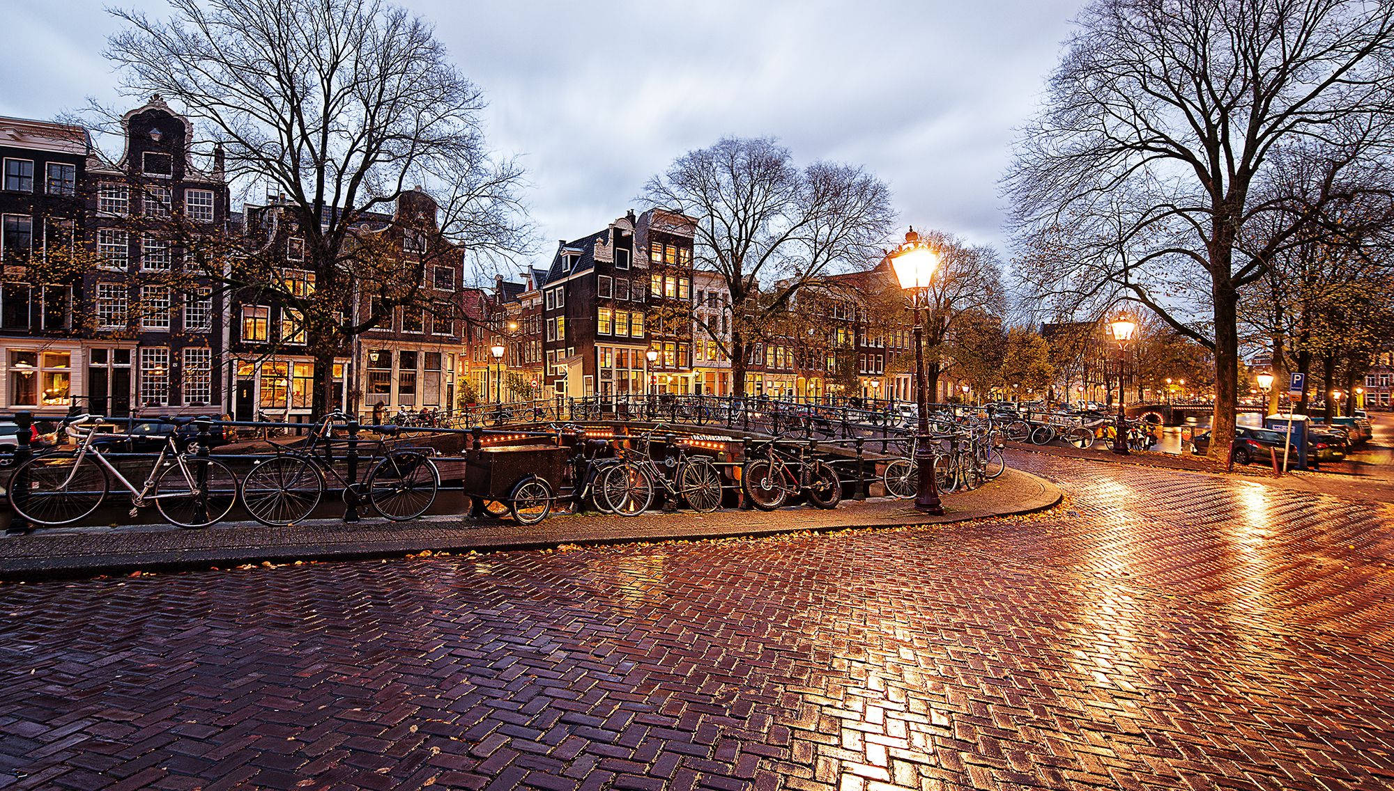 Netherlands 桌布 壁紙 — 阿姆斯特丹黃昏時分的濕漉漉鵝卵石運河廣場，發光的街燈和沿歷史建築停放的自行車。