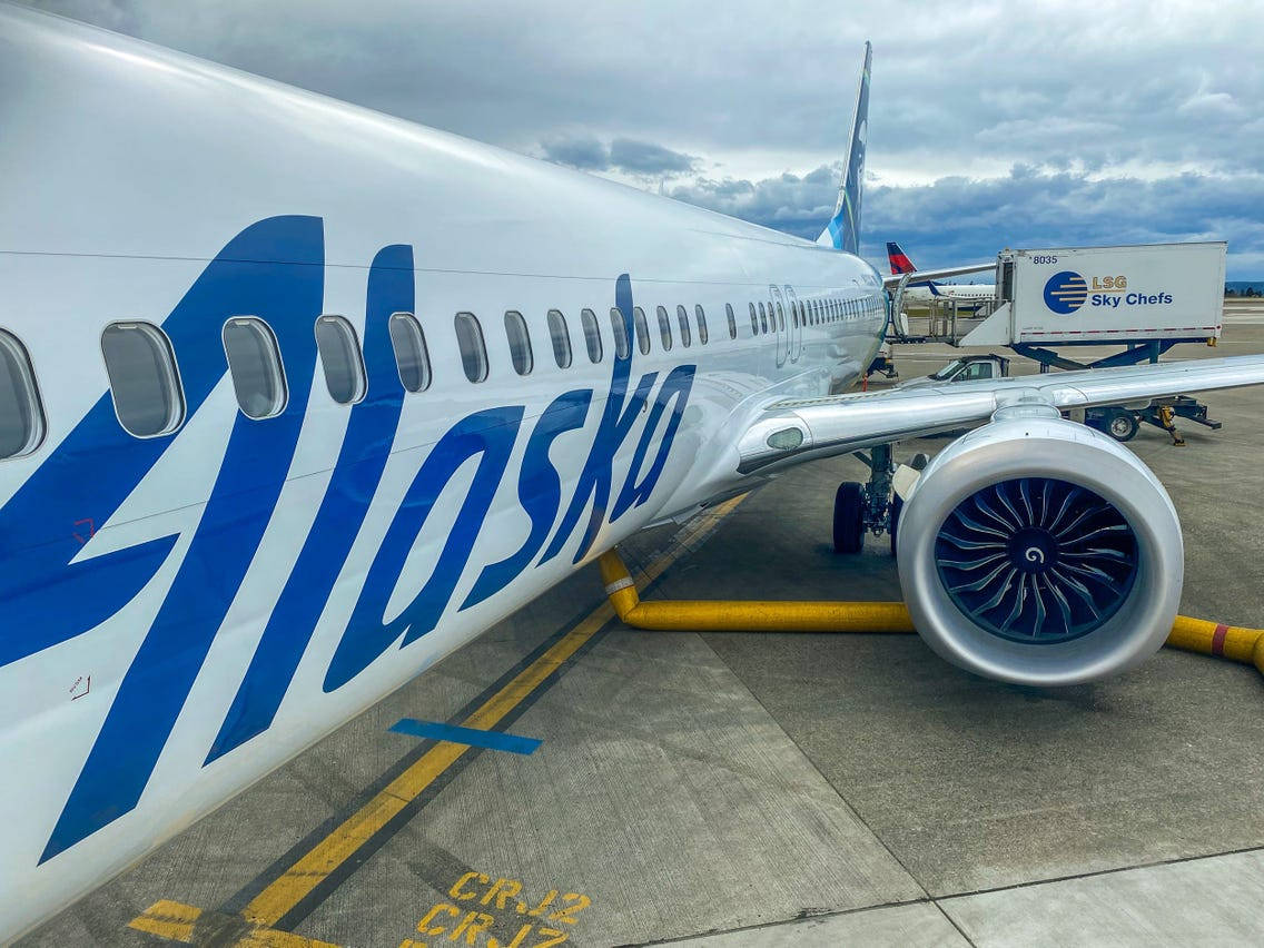 飛機 桌布 壁紙 — 阿拉斯加航空公司噴射機停泊在機場閘口，附近有貨車，天空多雲的特寫。