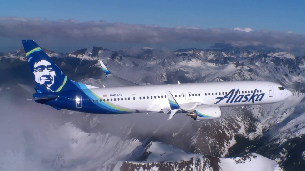  壁紙 — 阿拉斯加航空的飛機在晴朗藍天和零星白雲下，翱翔於積雪覆蓋的山脈之上。
