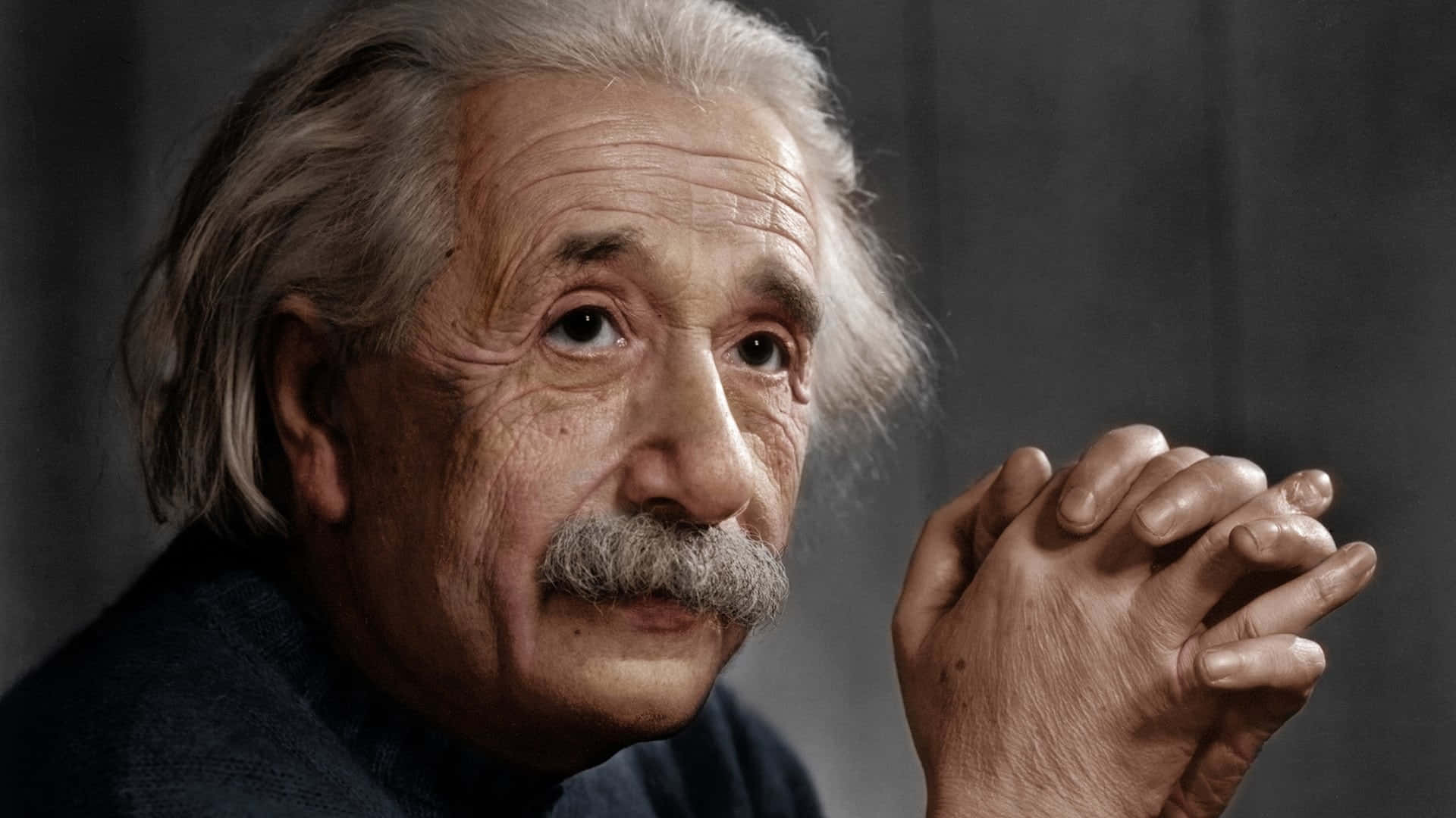 Albert Einstein 桌布 壁紙 — 阿爾伯特·愛因斯坦的頭像特寫，雙手合攏，目光沉思，背景為深灰色。