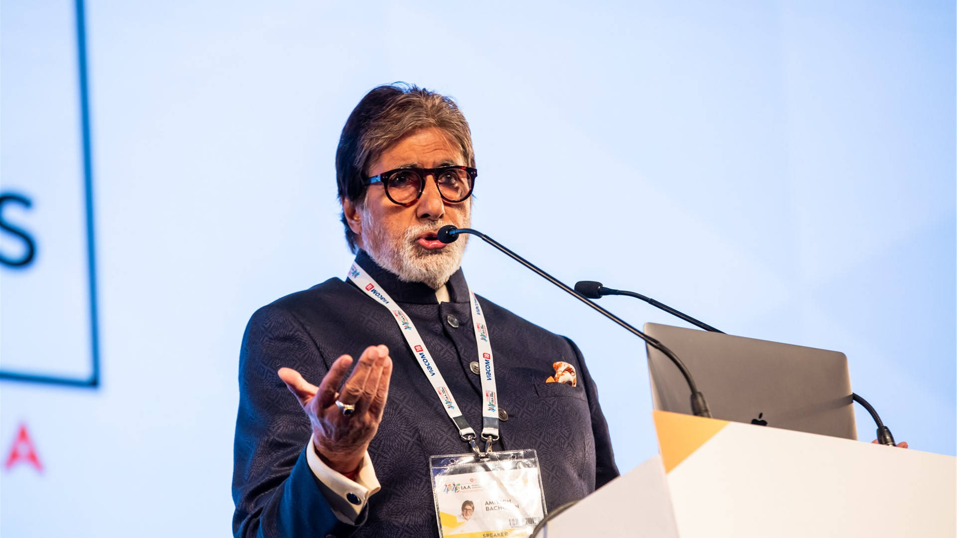 Amitabh Bachchan 桌布 壁紙 — 阿米塔布·巴強戴著眼鏡和深色圖案西裝，掛著證件帶，在會議上熱情演講。