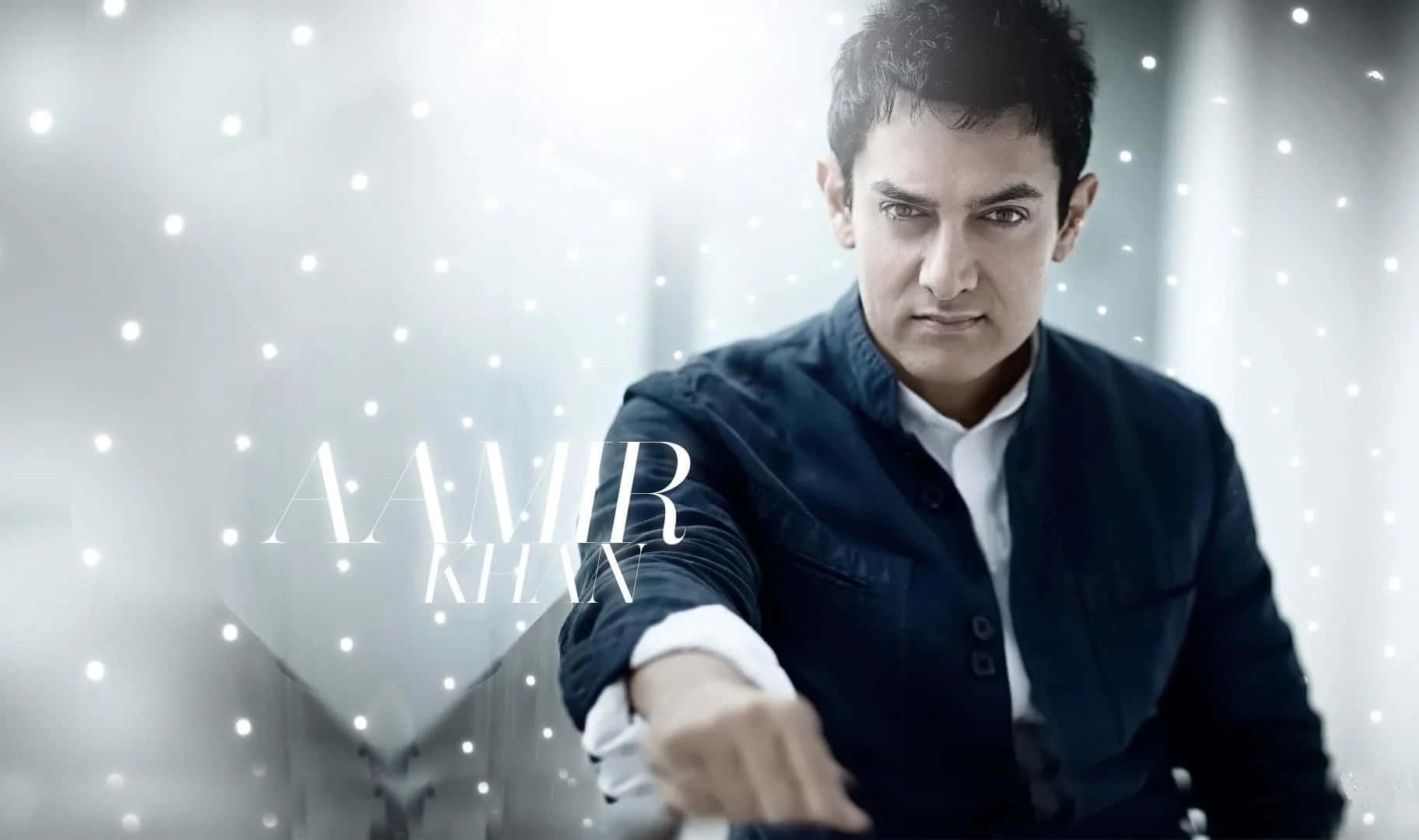 Aamir Khan 桌布 壁紙 — 阿米爾·汗身穿深色外套，在明亮閃爍的背景前伸出手，他的名字以優雅的白色文字呈現。