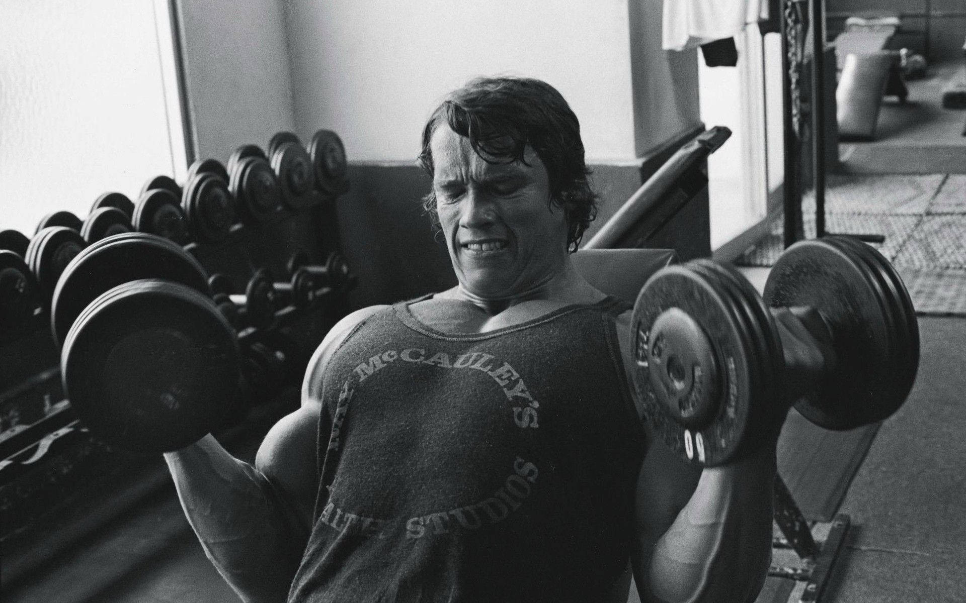 Arnold 桌布 壁紙 — 阿諾·史瓦辛格在粗糙的黑白健身房照片中，用力舉起沉重的啞鈴，表情緊張。