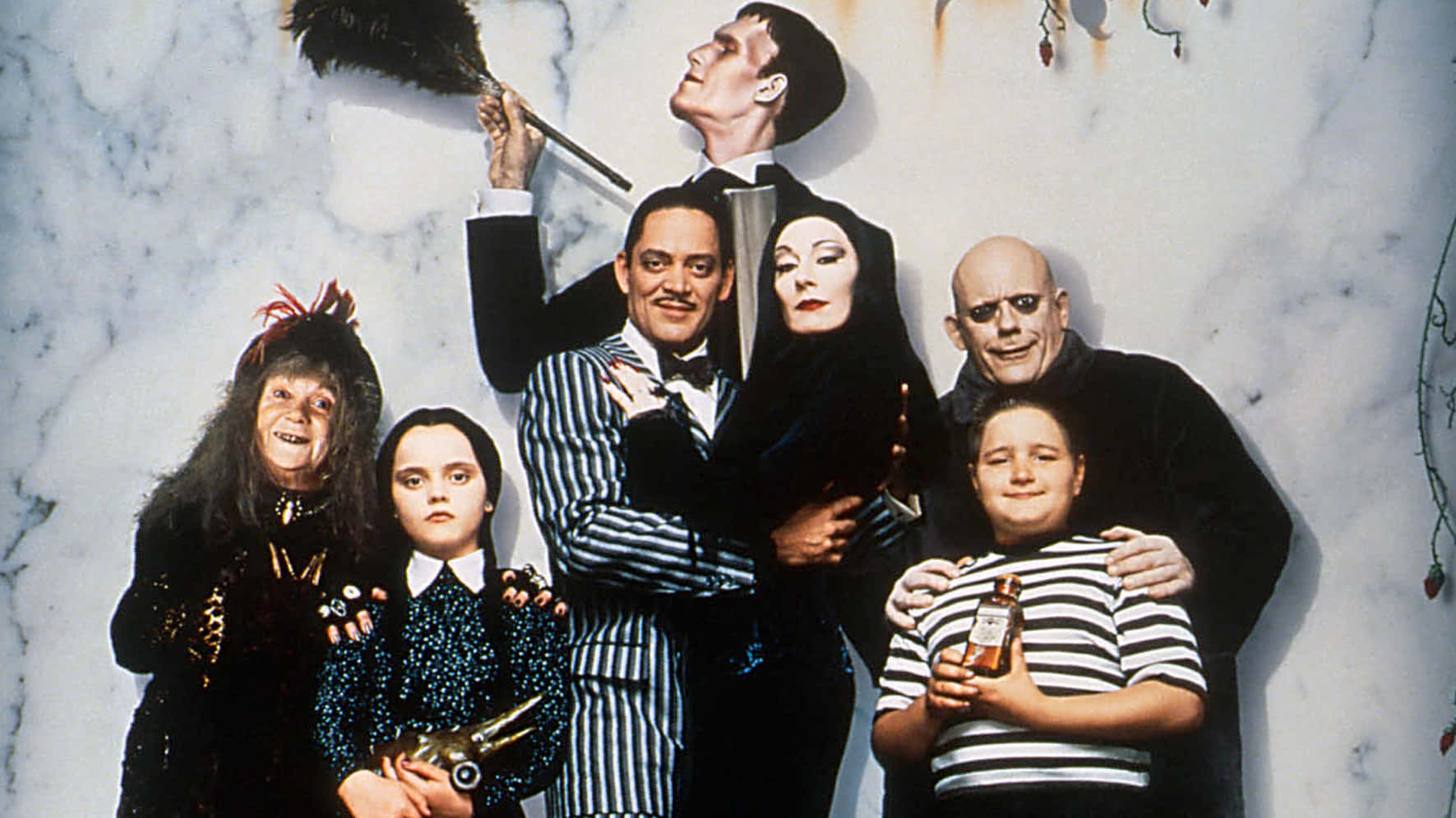 Addams Family 桌布 壁紙 — 阿達一族在大理石牆前戲劇性地擺姿勢，手持羽毛撣子和懸掛的裝飾品。
