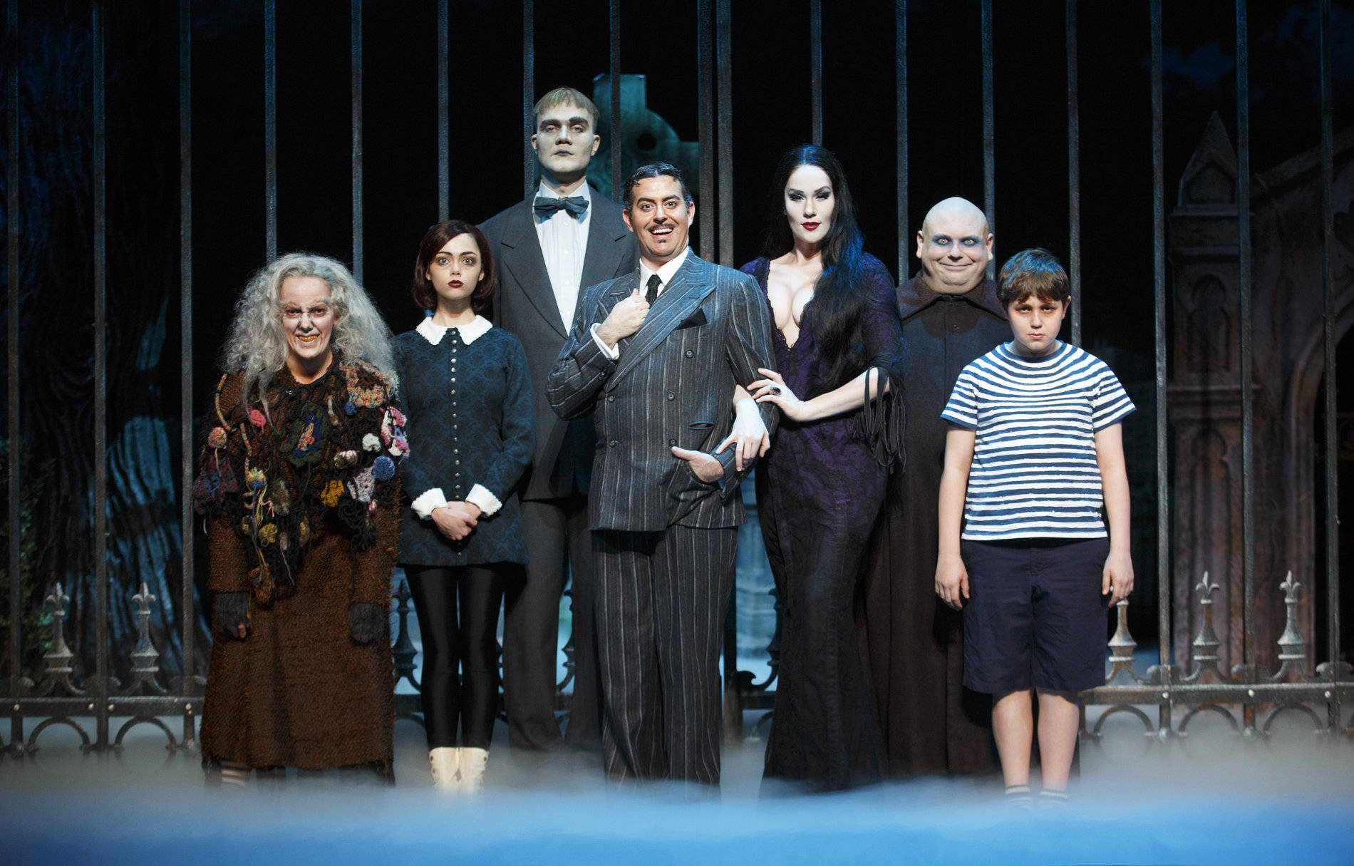 The Addams Family 桌布 壁紙 — 阿達一族演員以哥德式服裝戲劇性地擺姿勢，背景是黑暗的鐵門，腳邊有霧氣。