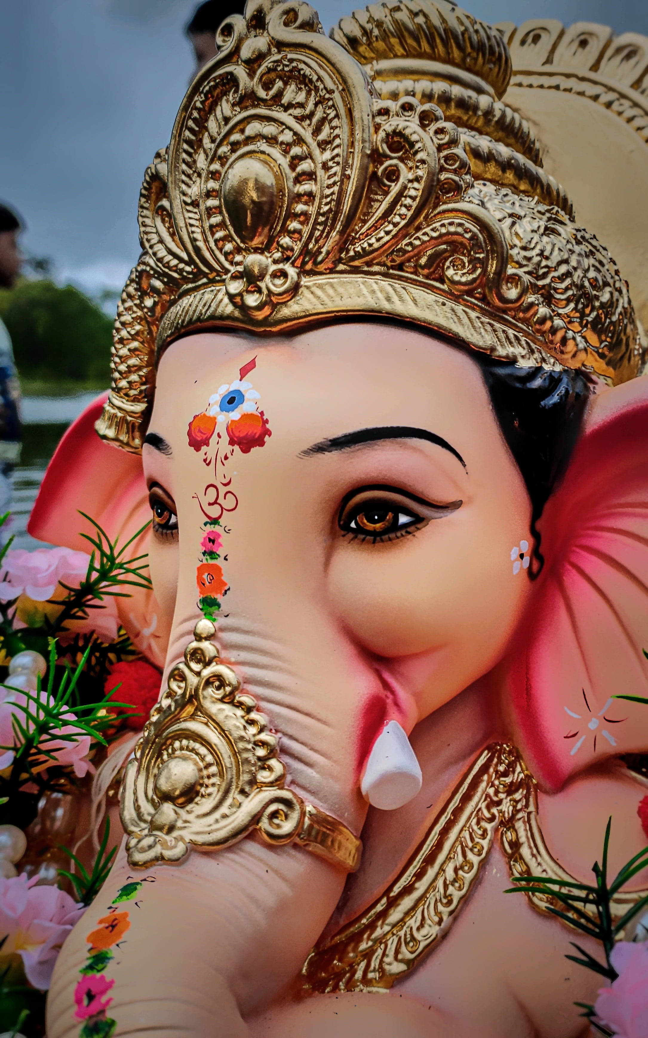 Ganesh 桌布 壁紙 — 陰天下一尊飾有金色珠寶和色彩繽紛花卉裝飾的鮮豔粉紅色象神雕像特寫。