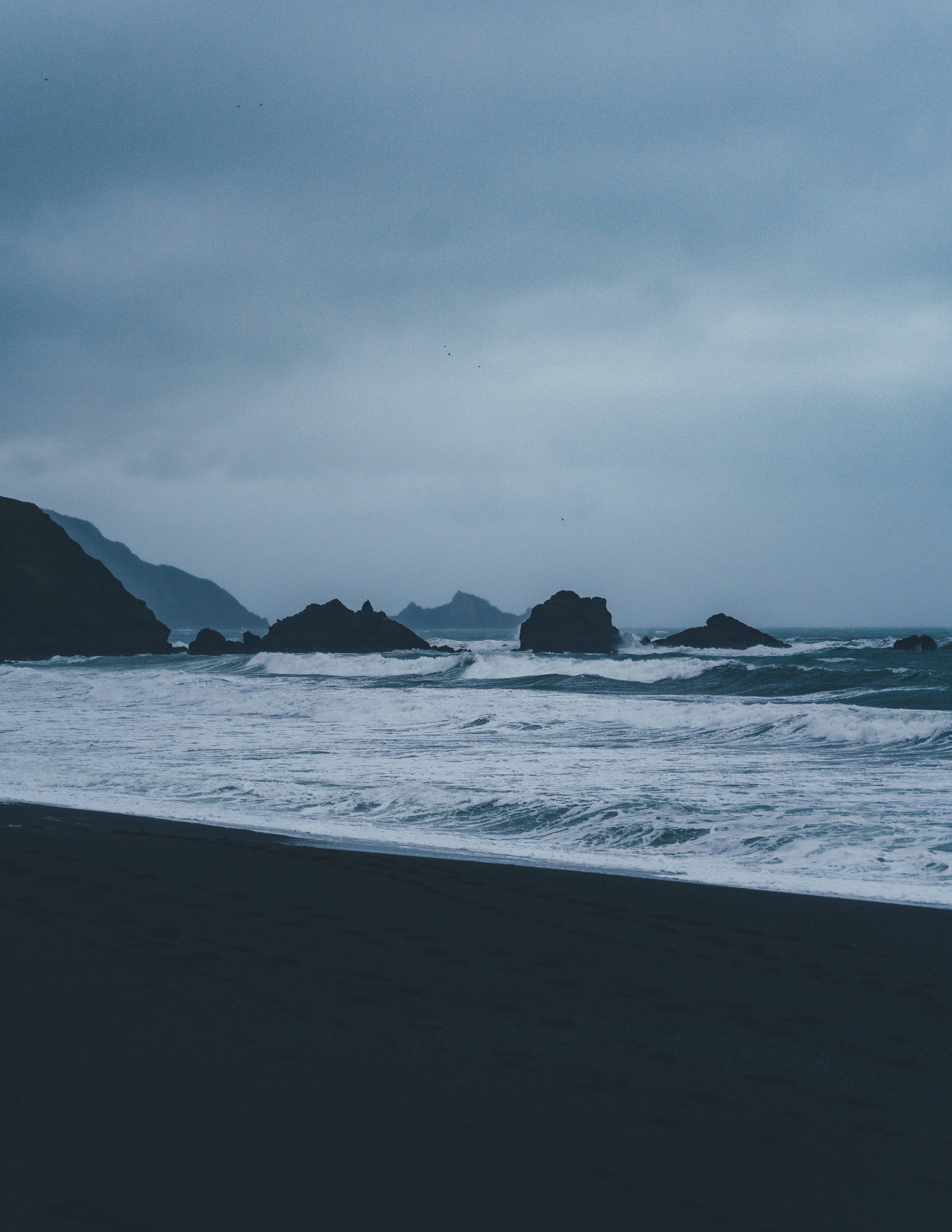 海洋 桌布 壁紙 — 陰天下一幅陰鬱的海岸景象，有深色火山沙、拍岸浪濤和參差不齊的岩石。