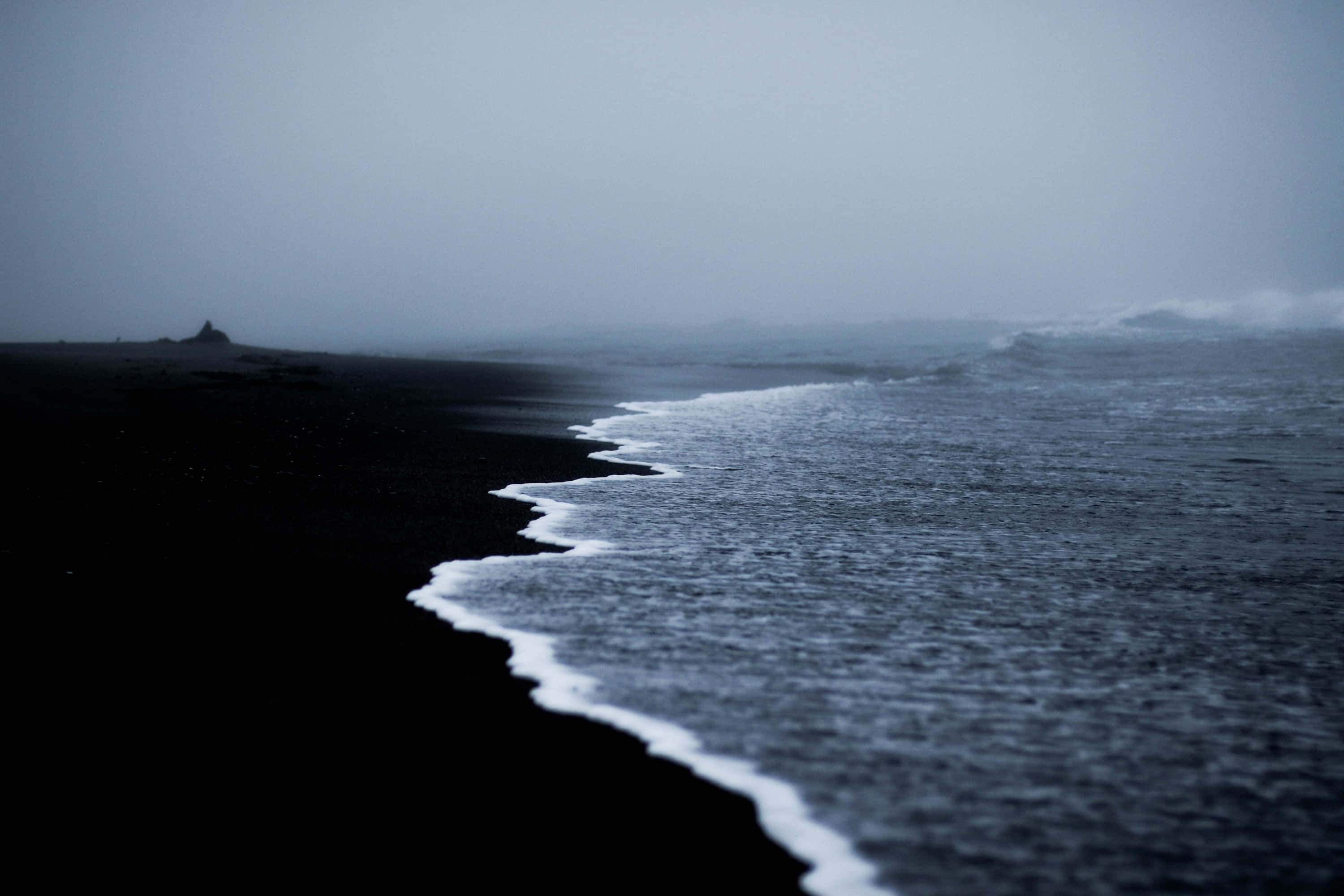 Black Sand Beach 桌布 壁紙 — 陰天下一幅陰鬱的海岸景象，深色火山沙與泡沫海浪交匯。