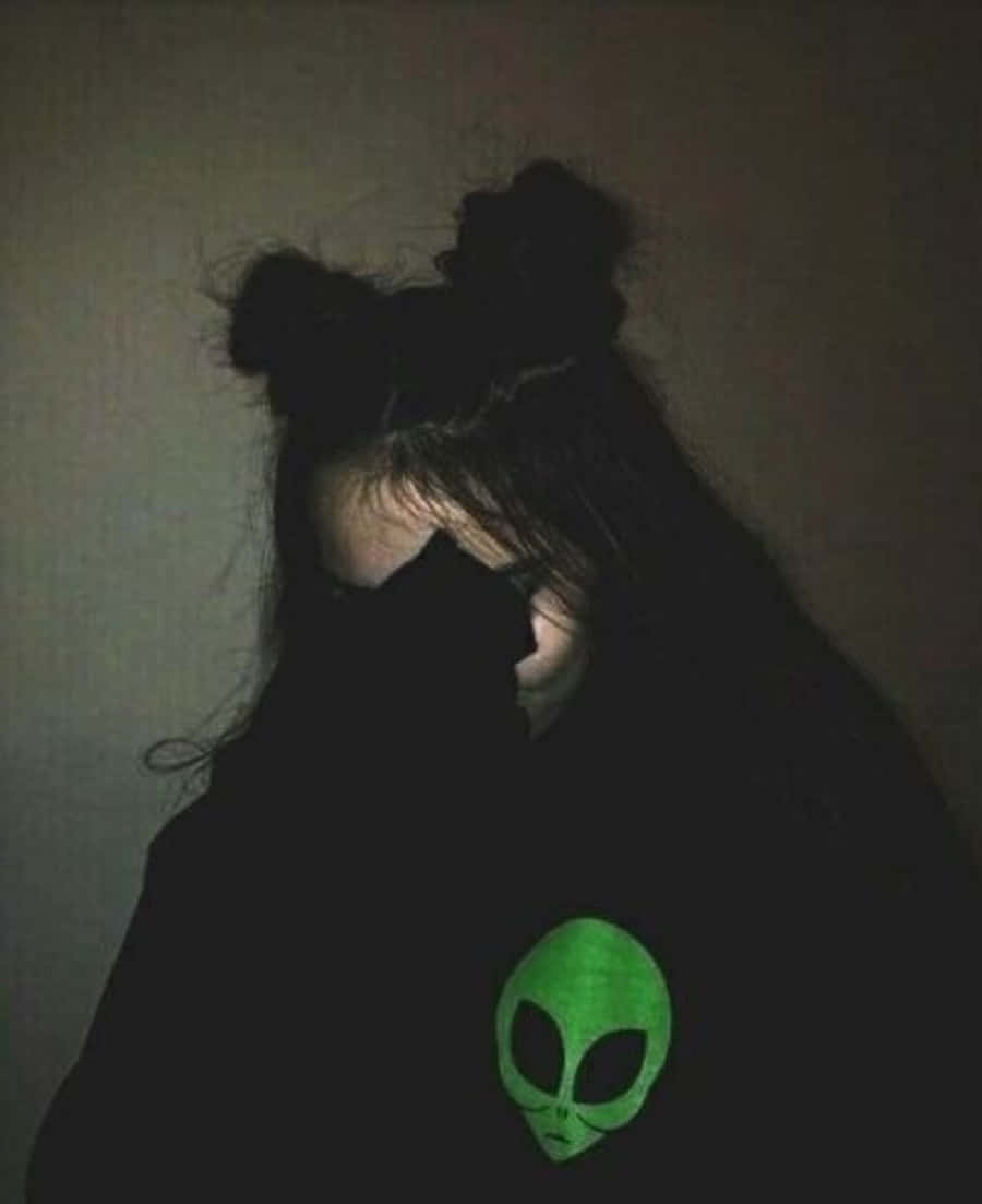 油漬搖滾 桌布 壁紙 — 陰影中的人穿著黑色連帽衫，胸前有一個發光的綠色外星人頭，背景是昏暗的牆壁。