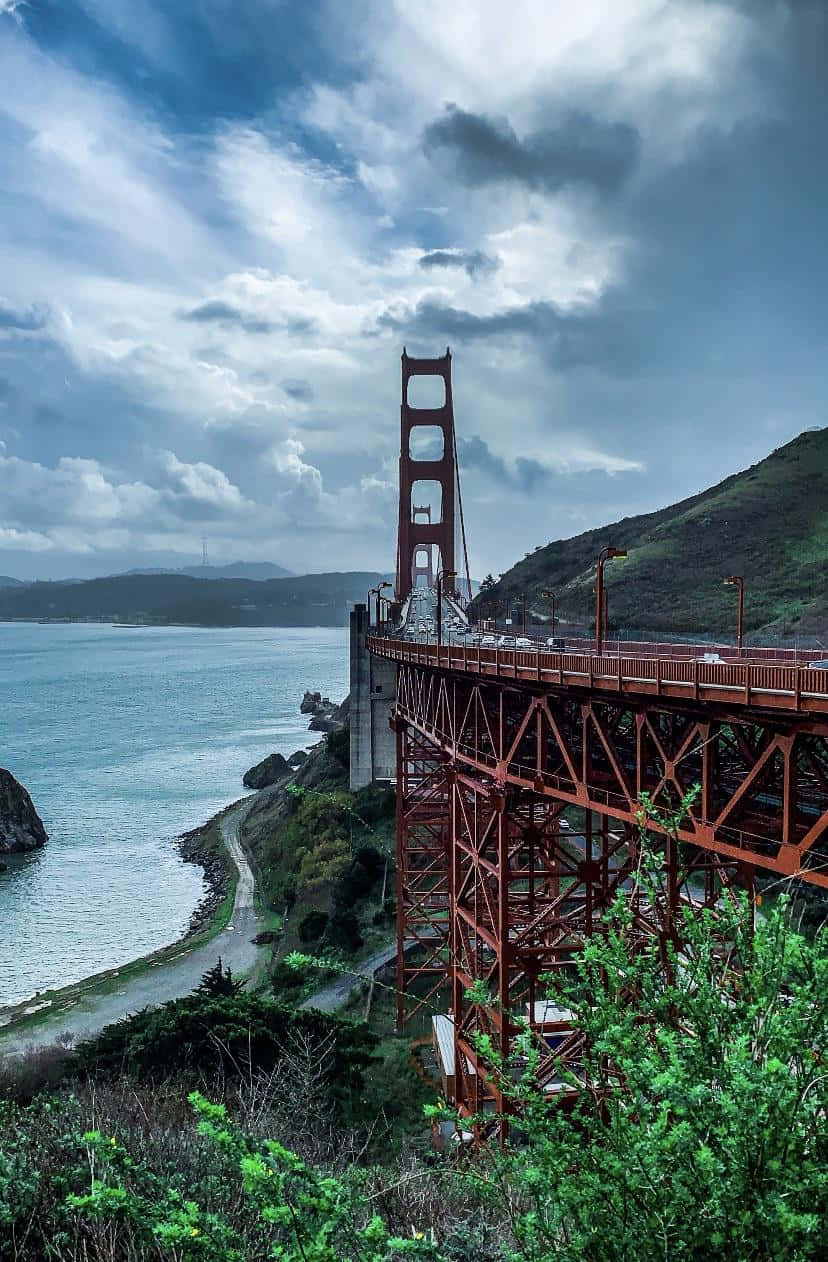 bridge 桌布 壁紙 — 陰沉多雲天空下金門大橋的戲劇性低角度景觀，配以茂密綠丘和藍色海水。