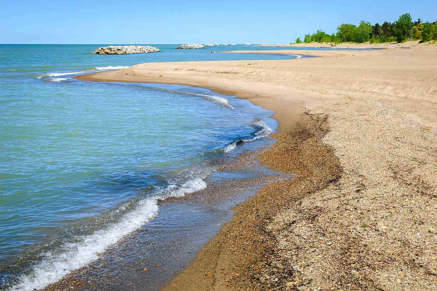 Lake Erie 桌布 壁紙 — 陽光下的海浪輕輕拍打著卵石海灘，彎向遠處樹木成蔭的海岸，天空清澈湛藍。