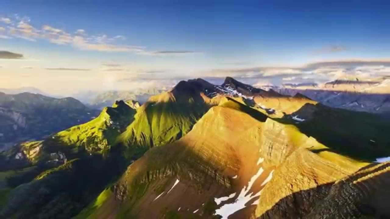 山景 桌布 壁紙 — 陽光以金色調沐浴崎嶇的山峰，雪塊依附在岩石斜坡上，廣闊的藍天之下。