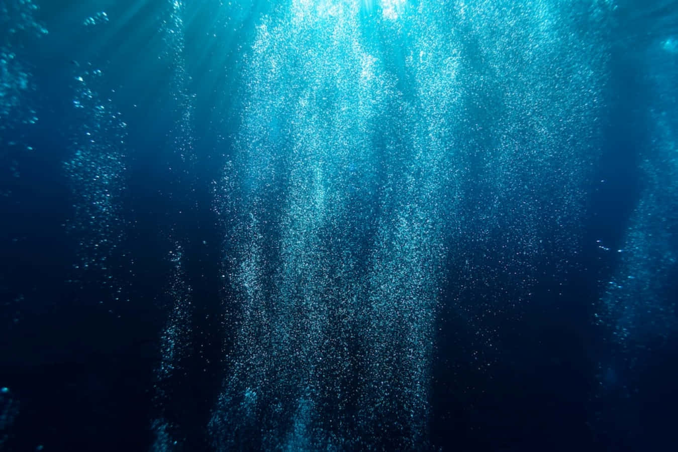 Deep 桌布 壁紙 — 陽光光束穿透深藍色海水，照亮向上升起的漩渦氣泡流。