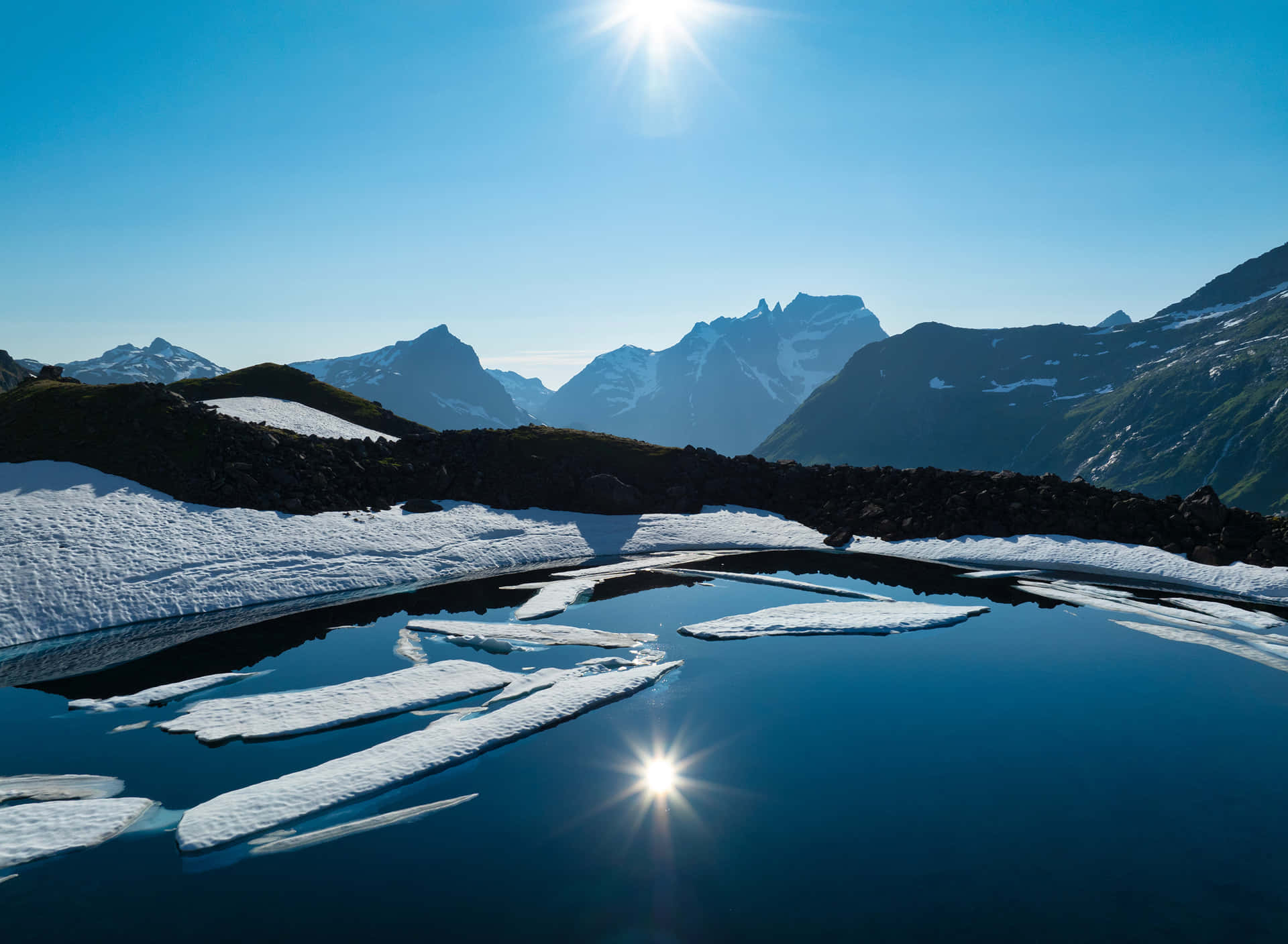 Lake View Reflection 壁紙 — 陽光在平靜的高山湖泊中漂浮的冰塊上閃爍，周圍是白雪覆蓋的山脈，天空清澈湛藍。