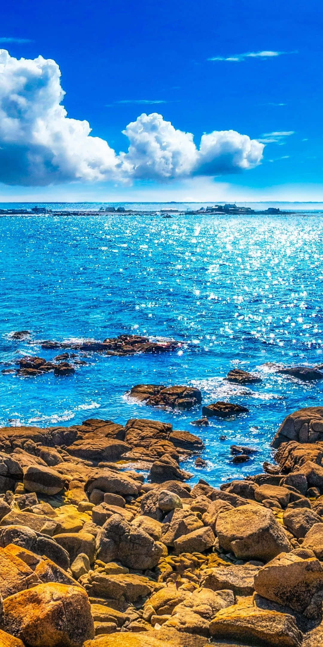 風景 桌布 壁紙 — 陽光在湛藍的海浪上舞動，拍打著金褐色的岩石海岸，天空湛藍，白雲朵朵。