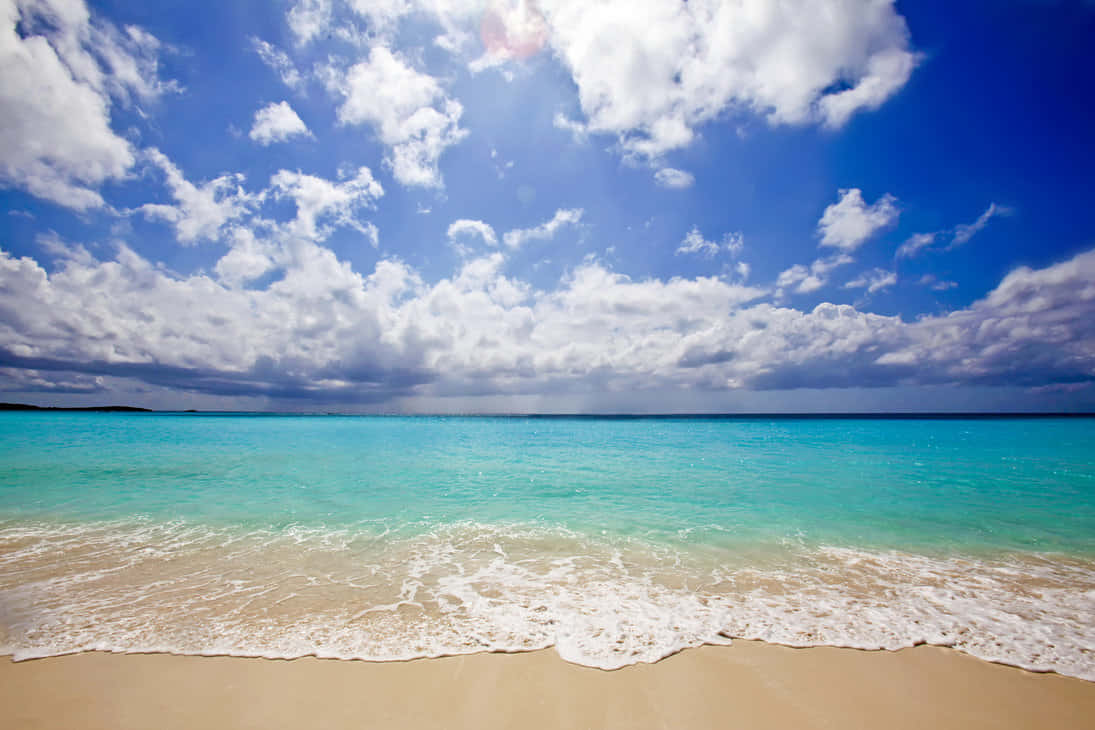 Caribbean Beach 桌布 壁紙 — 陽光在碧綠色的海浪上舞動，海浪輕拍著金色的沙灘，上方是廣闊的天空，滿佈蓬鬆的白雲。