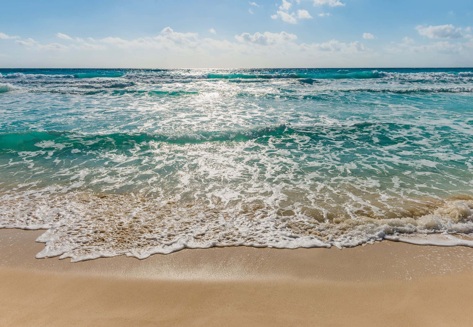 Beach Scene 桌布 壁紙 — 陽光在碧綠色的海浪上閃爍，拍打著金色的沙灘，天空湛藍，白雲點綴。