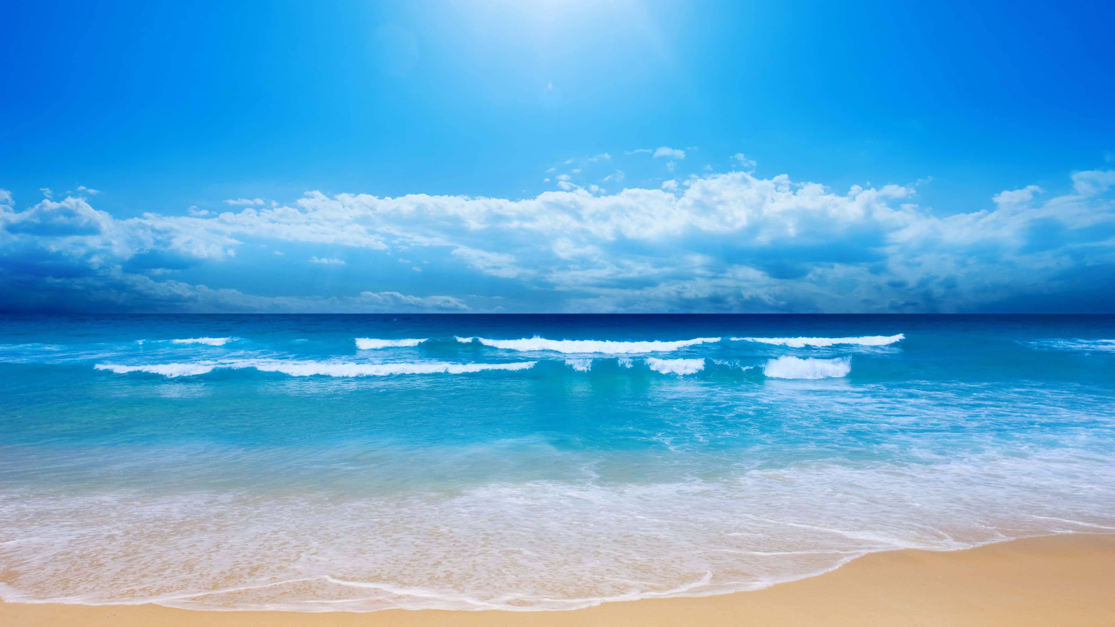 4K 海灘壁紙 桌布 壁紙 — 陽光在碧綠色的海浪上閃爍，拍打著金色的沙灘，頭頂是廣闊的藍天和蓬鬆的白雲。