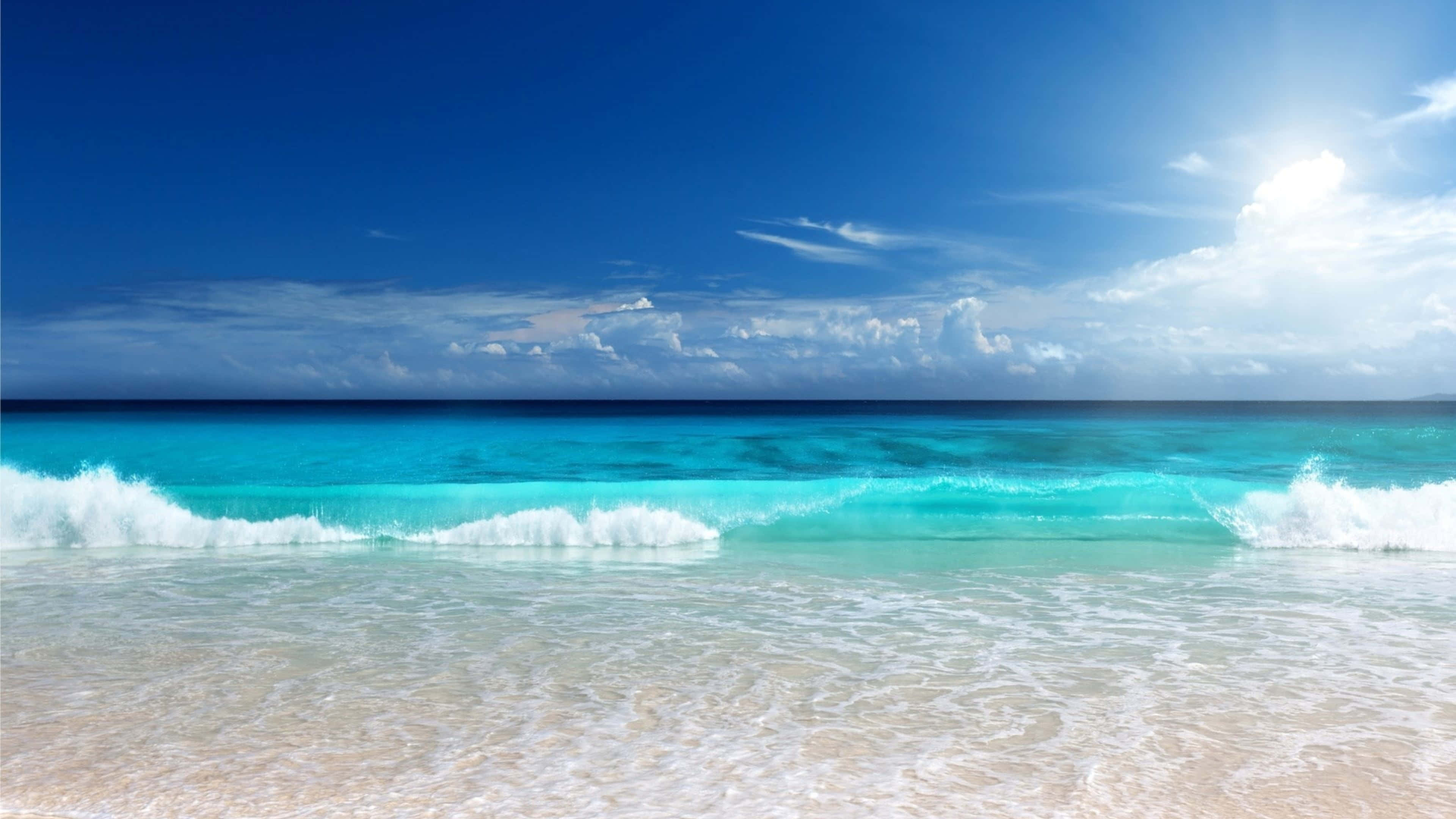 Beautiful Beach 桌布 壁紙 — 陽光在碧綠色的海浪上閃爍，海浪拍打著純淨的白色沙灘，上方是廣闊的藍天和零星的白雲。