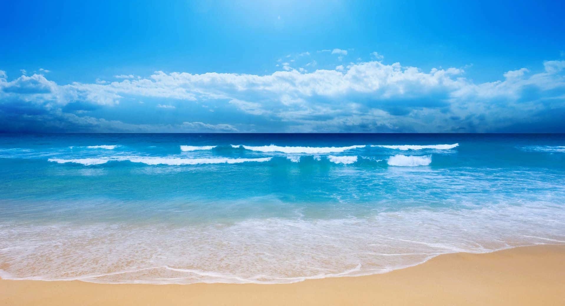 海灘 桌布 壁紙 — 陽光在碧綠色的海浪上閃爍，海浪拍打著金色的沙灘，上方是廣闊的藍天和蓬鬆的白雲。