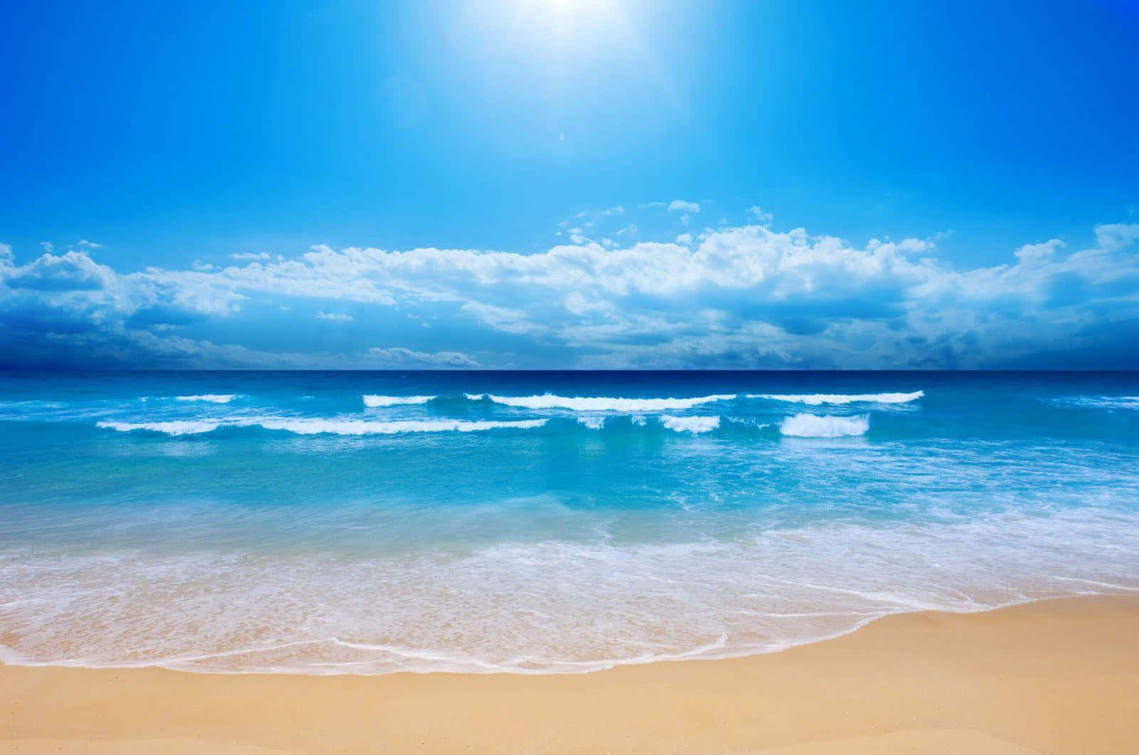 Beach Desktop 桌布 壁紙 — 陽光在蔚藍的海洋上閃耀，泡沫般的波浪在部分多雲的天空下滾滾湧上金色沙灘。