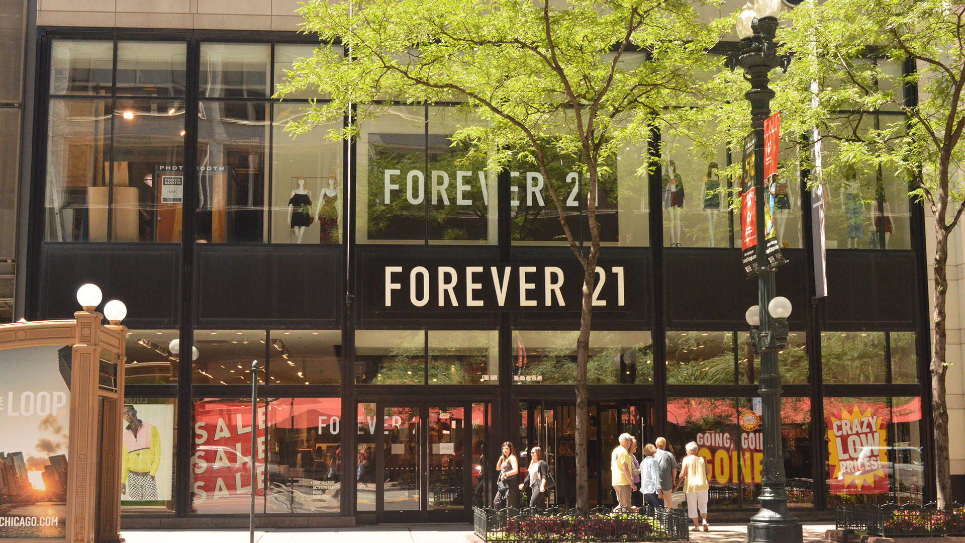 永遠21 桌布 壁紙 — 陽光明媚的城市街景，Forever 21 商店的玻璃櫥窗展示著人體模型和促銷標誌，周圍環繞著綠樹和行人。