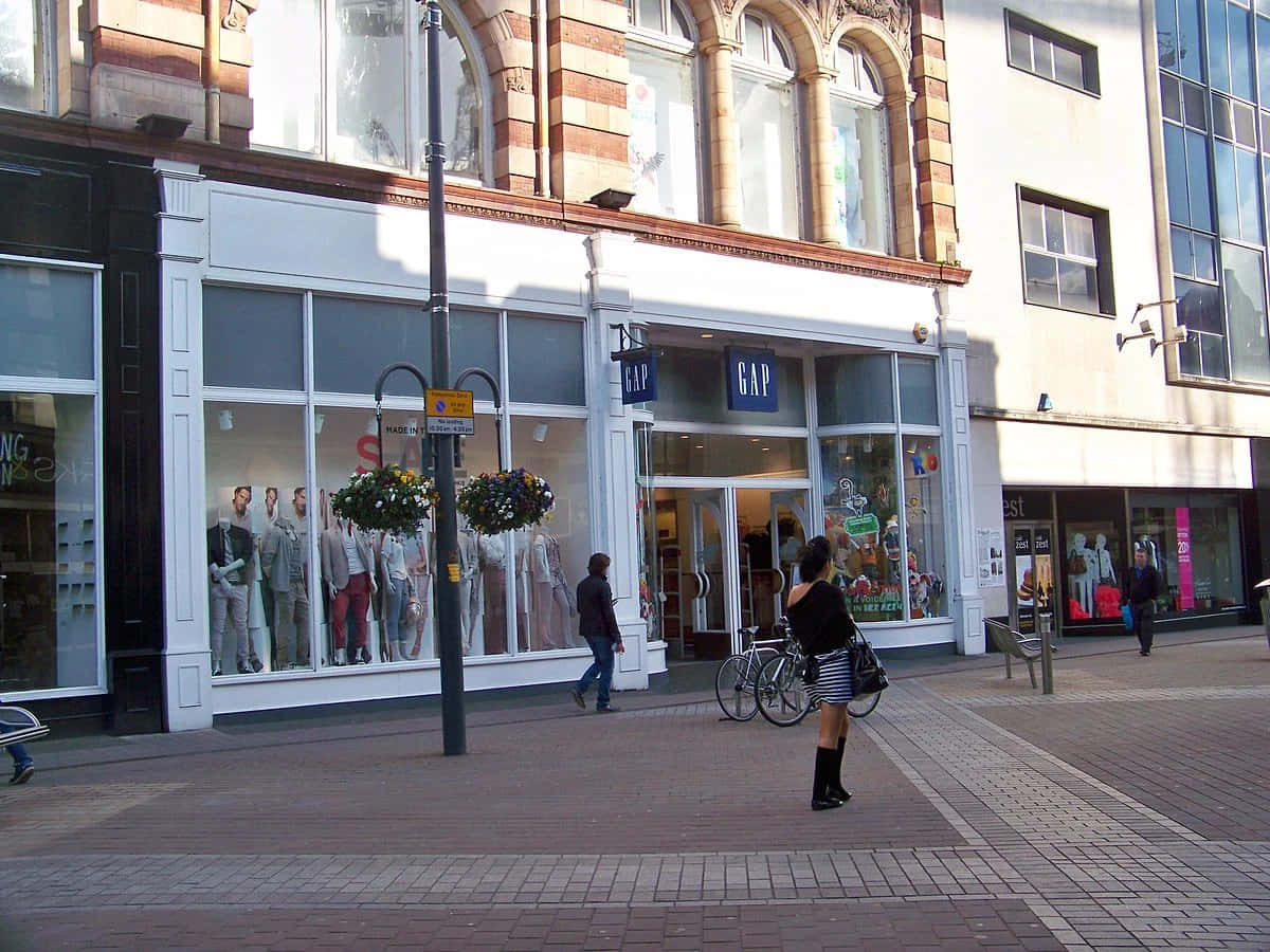 Gap 桌布 壁紙 — 陽光明媚的城市街景，GAP服裝店櫥窗裡有模特兒，行人走過磚砌人行道。