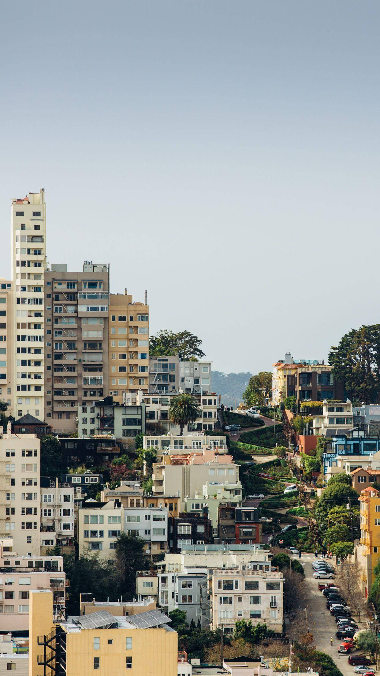 San Francisco Phone 桌布 壁紙 — 陽光明媚的山坡城市景觀，色彩繽紛的房屋沿著陡峭的斜坡層疊而下，朝向現代高樓大廈，背景是清澈的藍天。