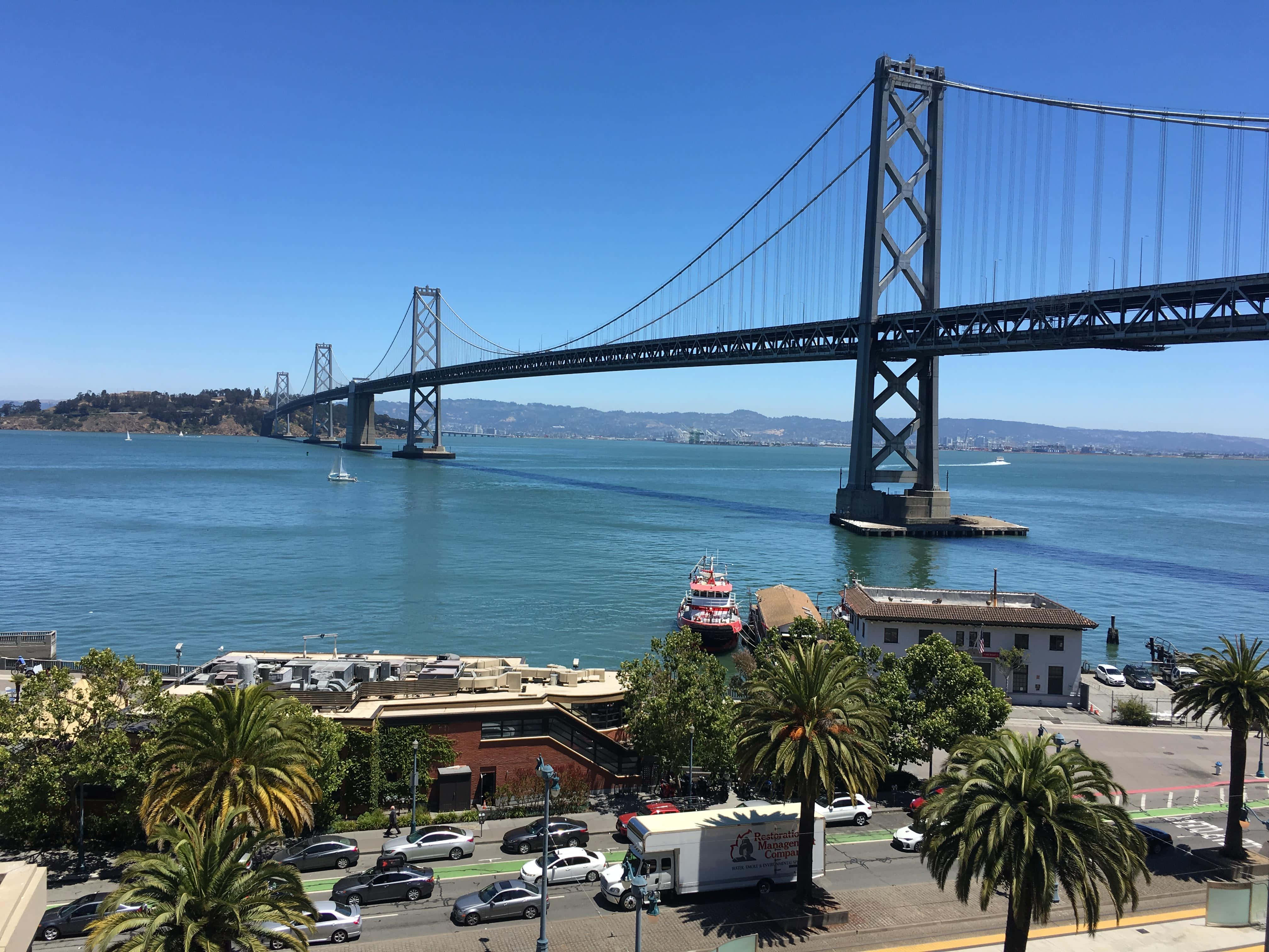Beautiful View 桌布 壁紙 — 陽光明媚的日子，海灣大橋橫跨碧綠海水，下方有棕櫚樹和城市街道。
