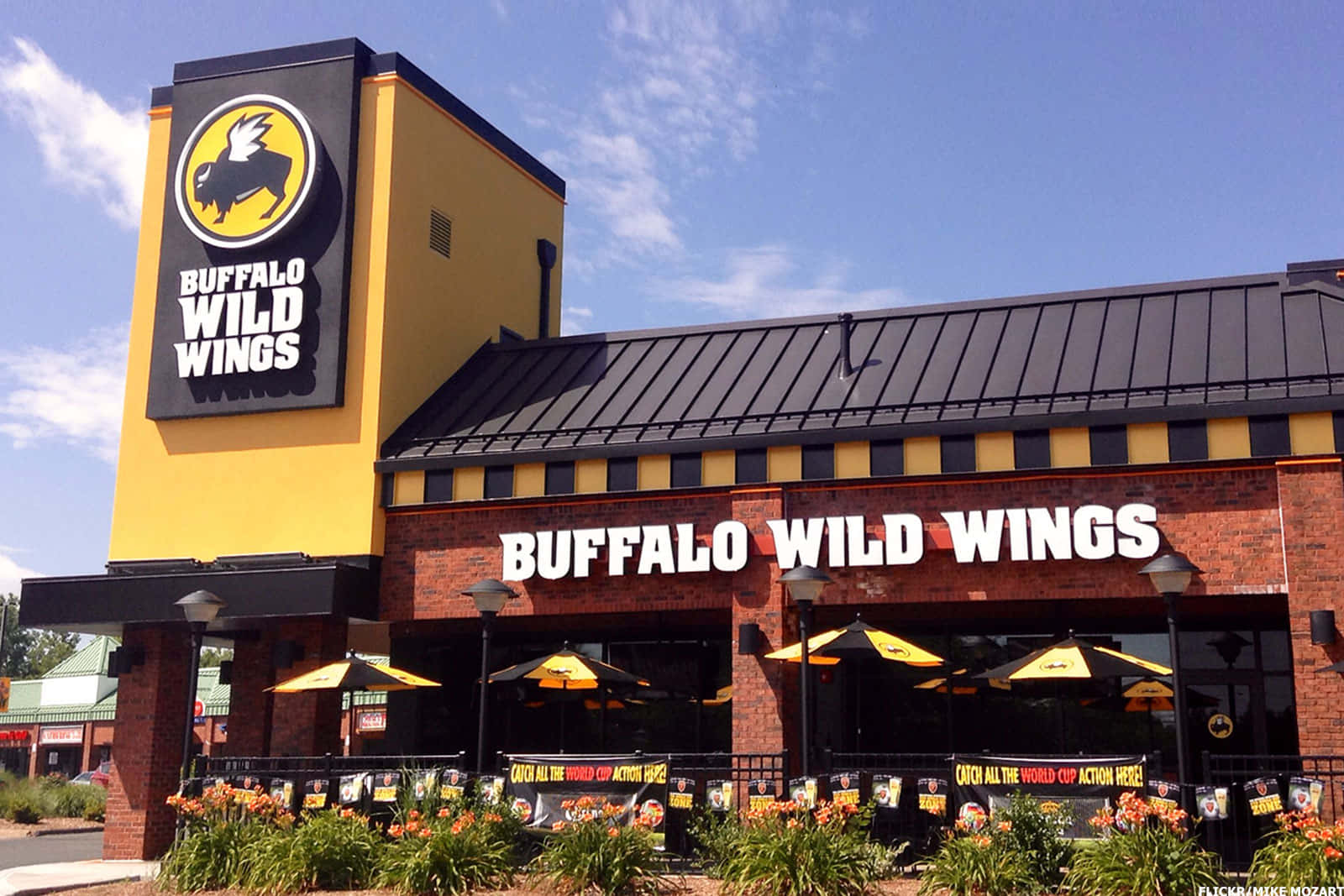水牛城辣雞翅 桌布 壁紙 — 陽光明媚的日子裡，一間明亮的黃色Buffalo Wild Wings餐廳，磚砌外牆，黃色陽傘下設有戶外座位。