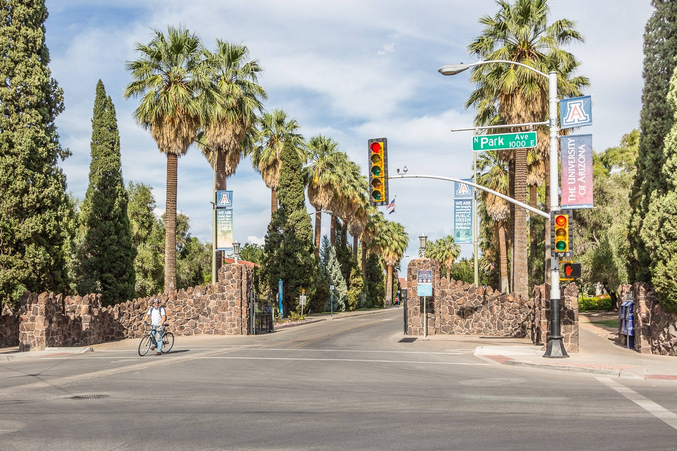 Arizona 桌布 壁紙 — 陽光明媚的校園入口，有棕櫚樹、石牆和一名騎自行車者在交通燈下經過。